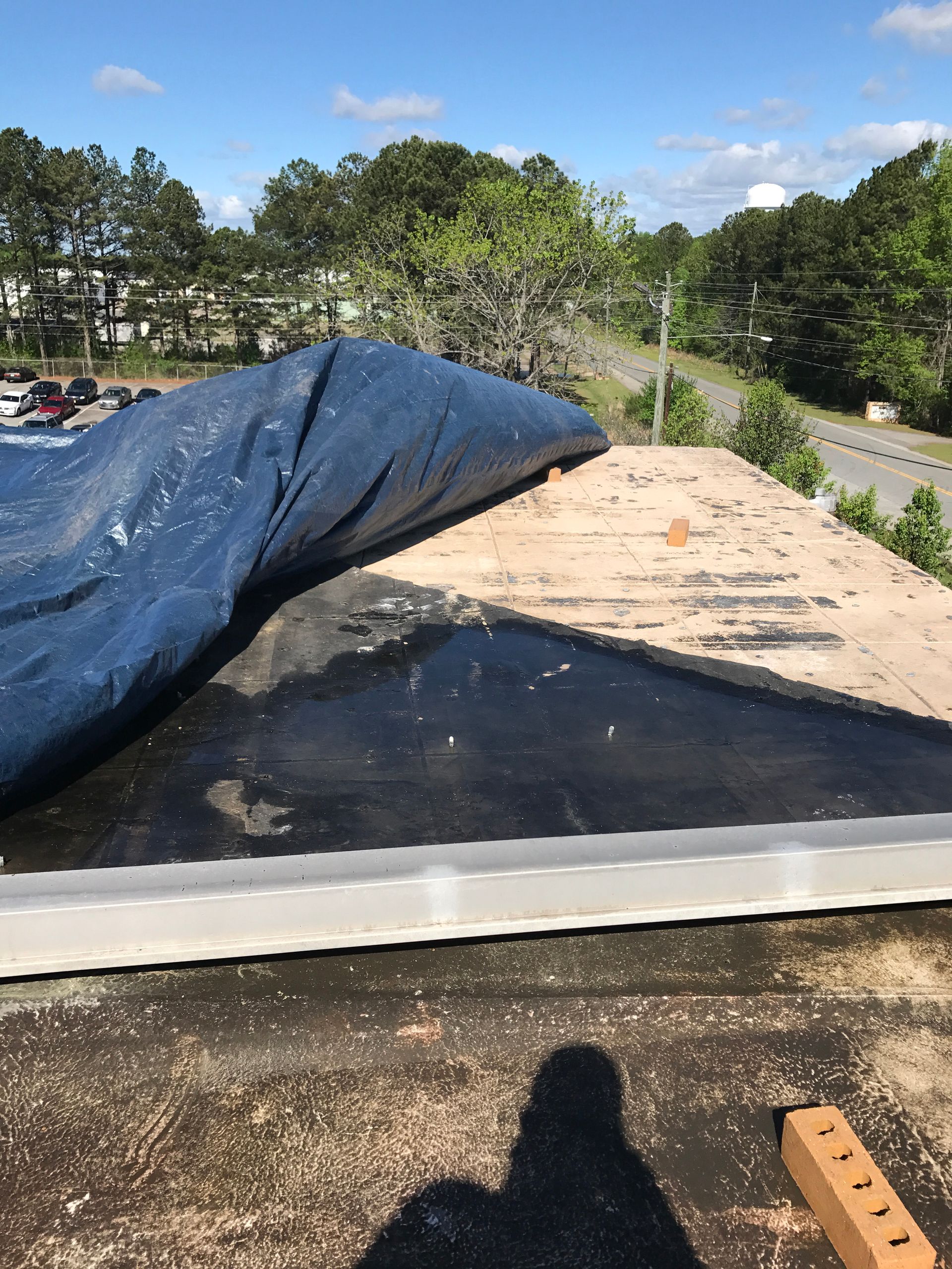 Blue tarp partially covers a weathered roof with scattered debris, viewed outdoors on a sunny day.