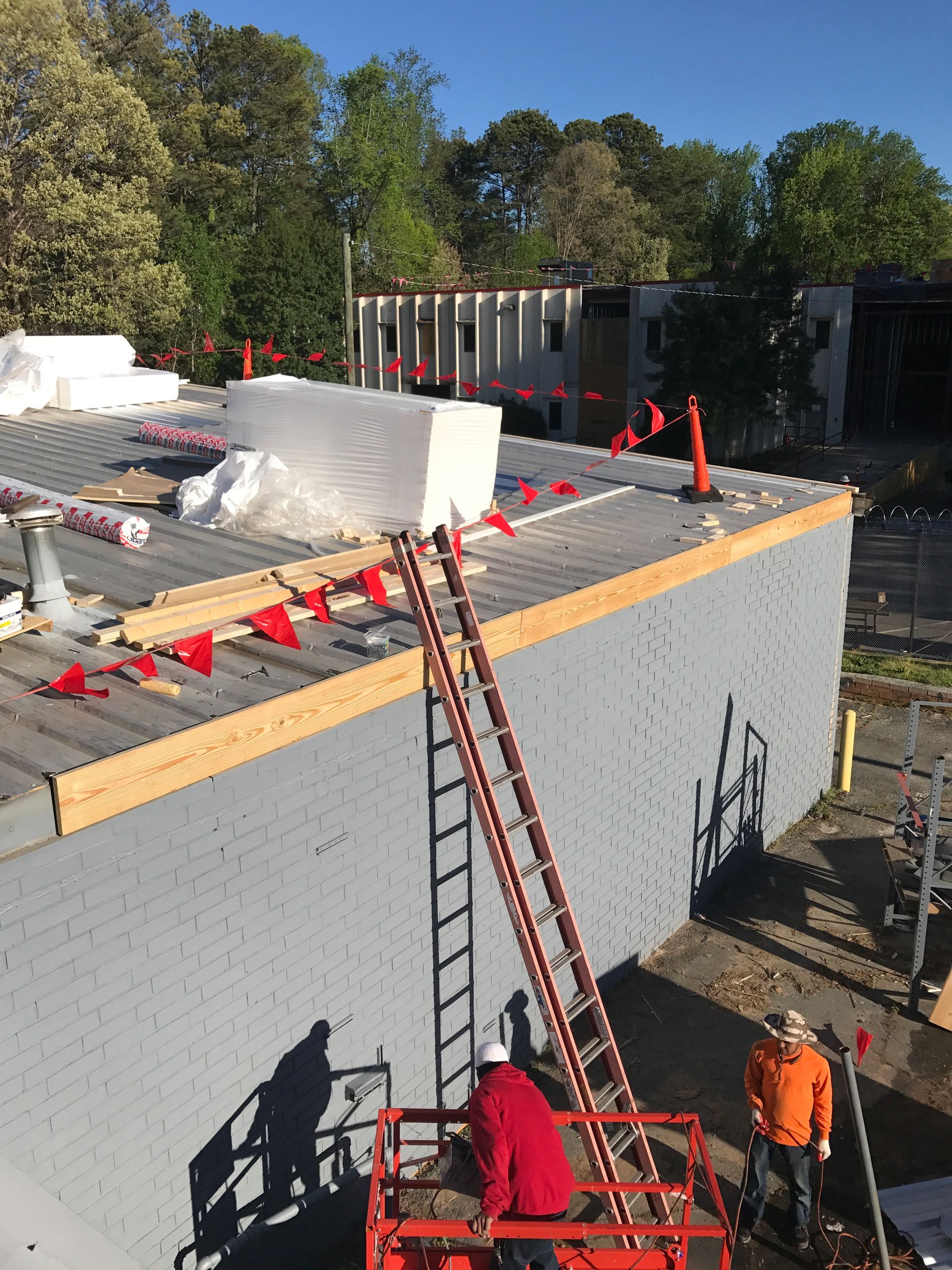 Construction workers on a roof, with a ladder and lift. Red flags line the edge.