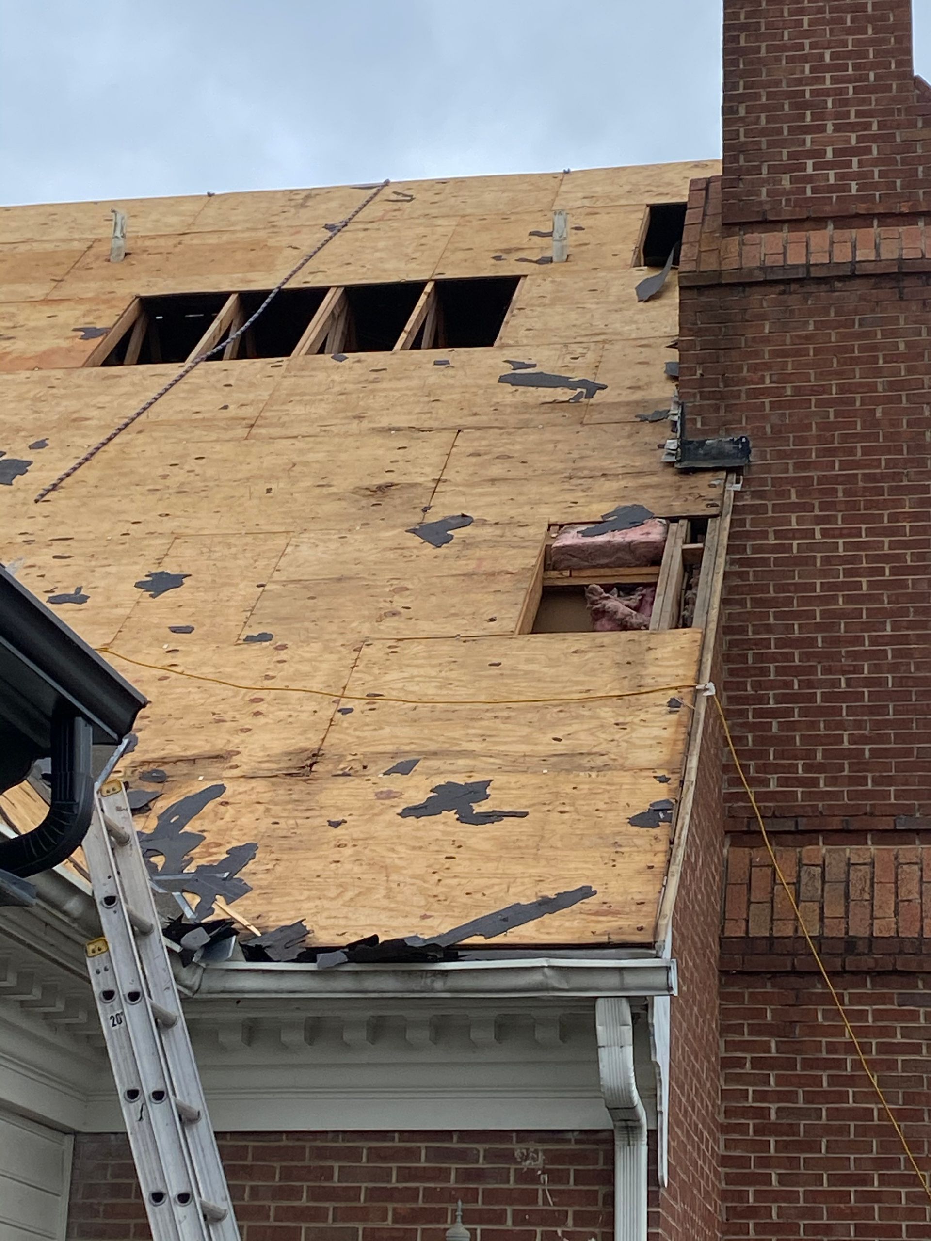 Partially torn-off roof of a brick house; missing shingles reveal plywood and insulation; a ladder leans against the gutter.