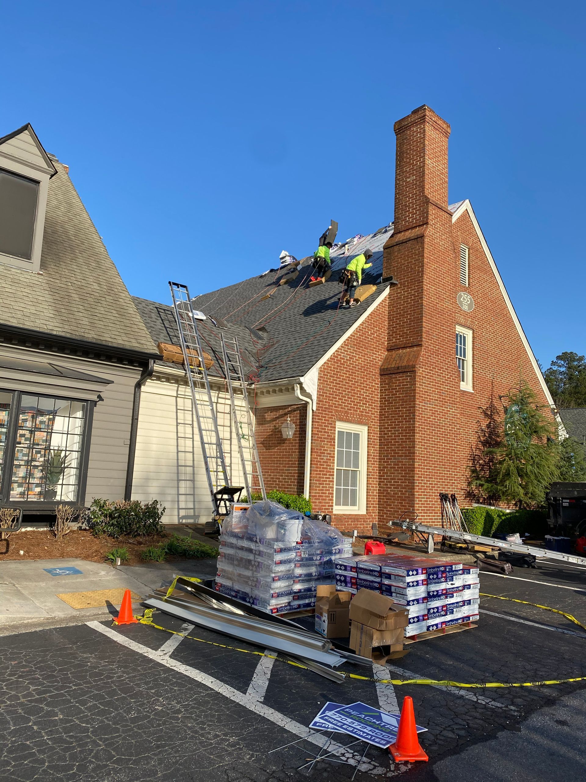 Roofers replacing a shingle roof on a brick building with a tall chimney. Equipment and materials are on the ground.