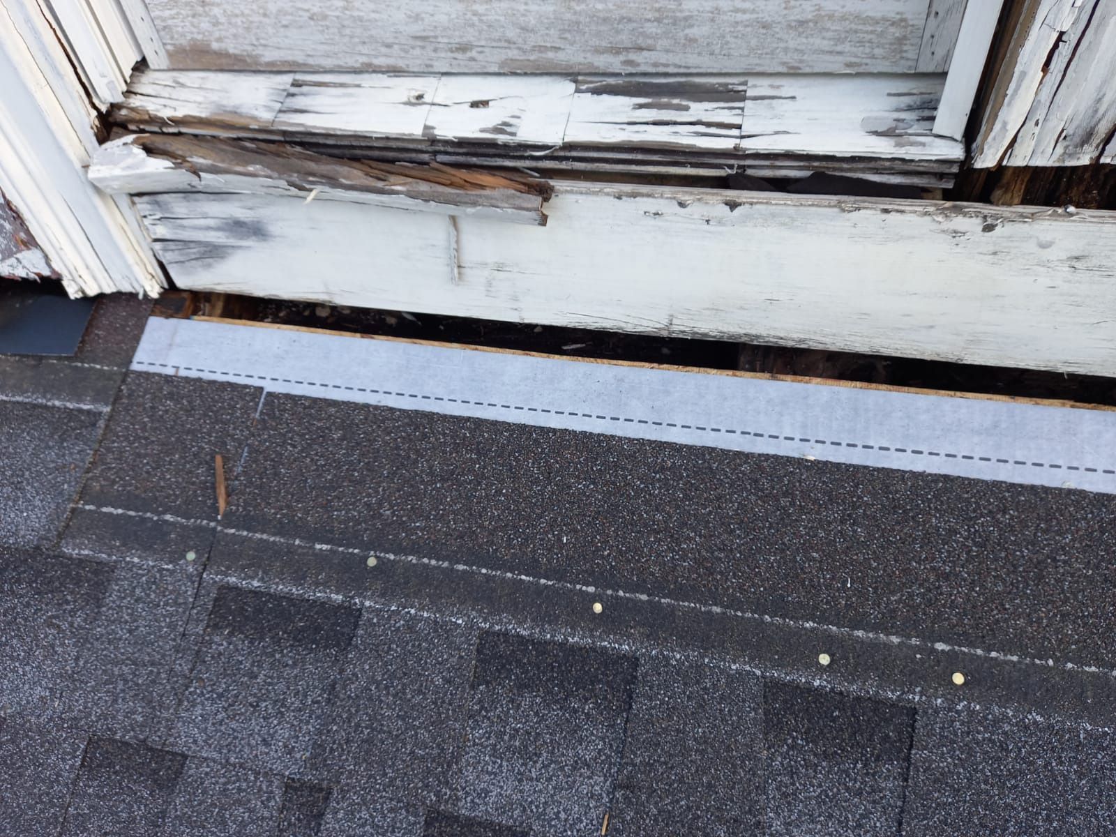 Damaged wooden trim above dark gray shingles. Flashing tape visible between.