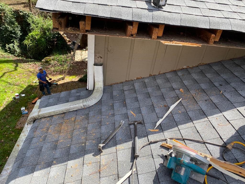 Roofer working on a roof section with missing wood. Tools and debris are scattered on the roof. Sunny day.