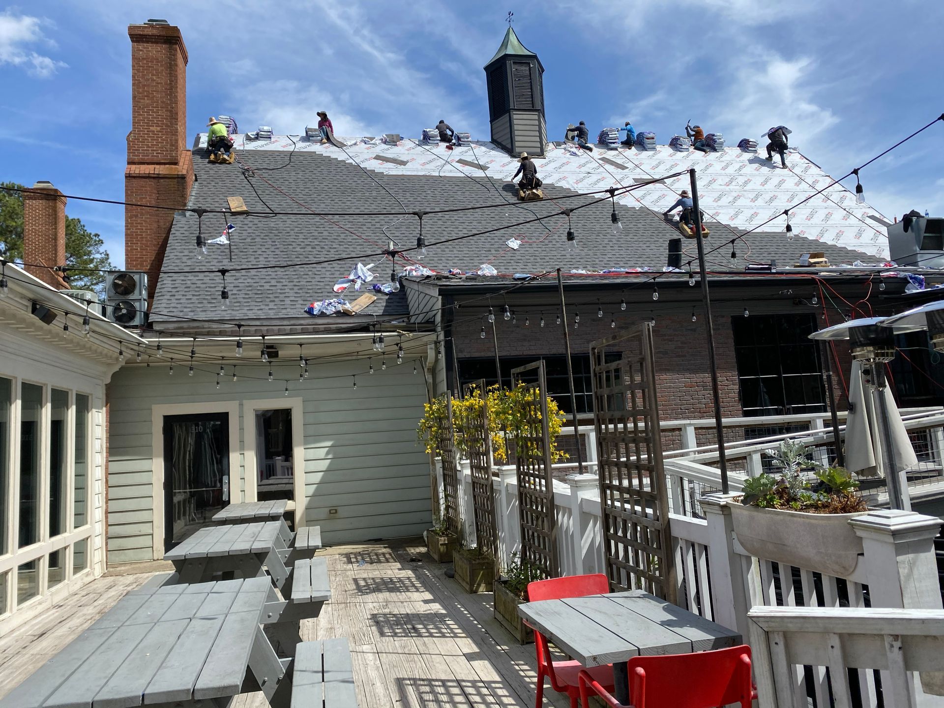 Roofing workers on a building with a brick chimney and a light-strung outdoor patio. Blue sky.