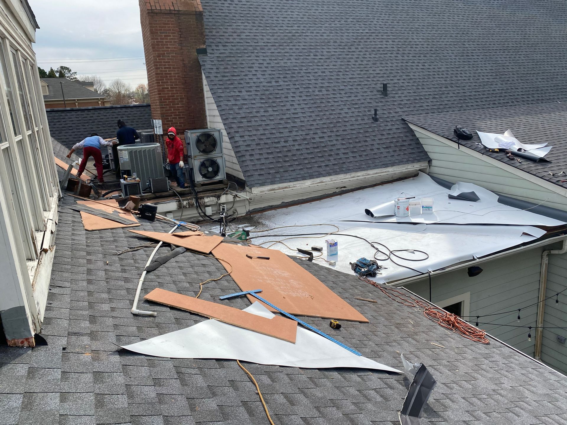 Roof repair in progress: Workers near air conditioning units, shingles, and building with chimney.