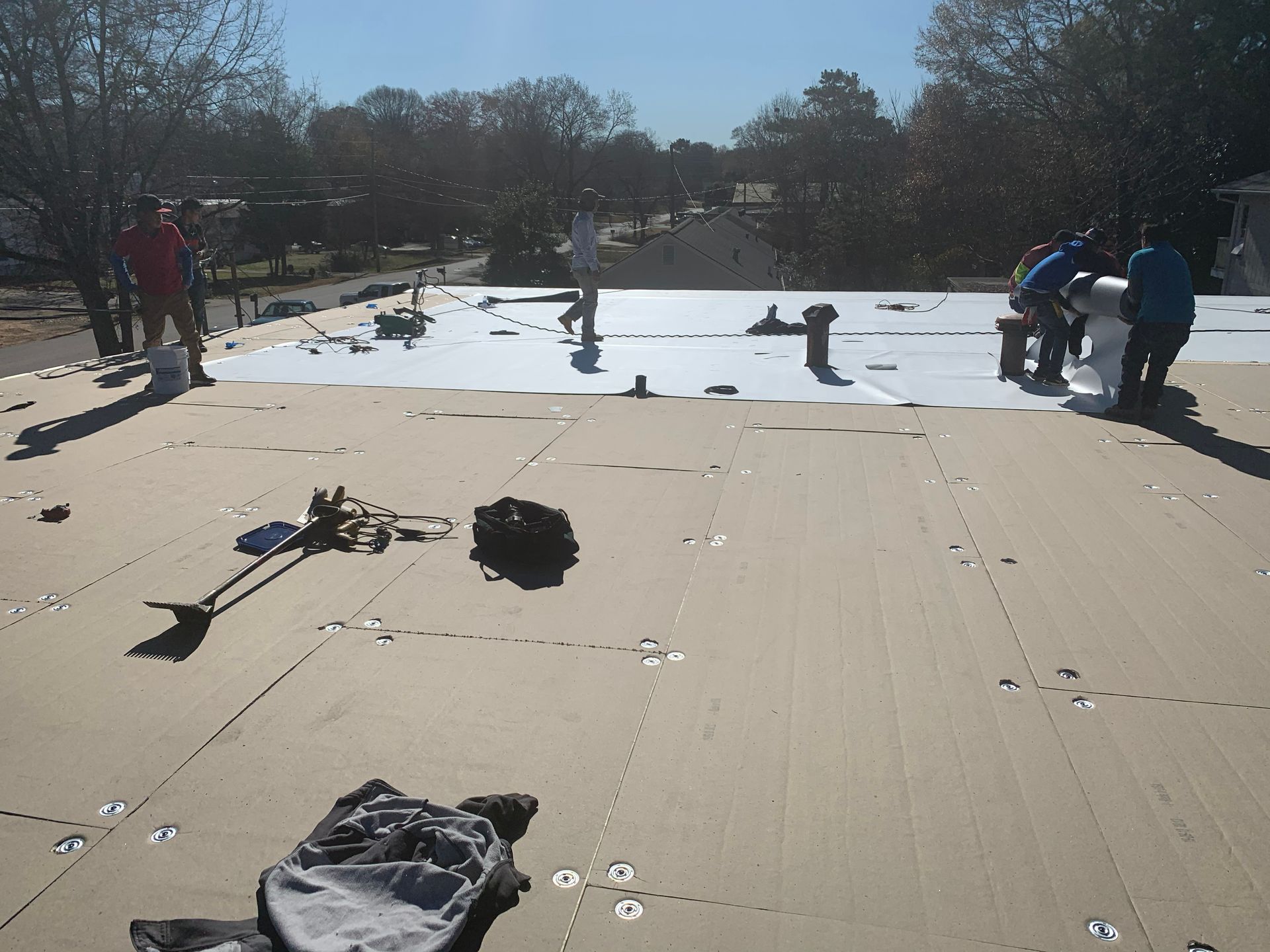 Workers on a flat roof installing white roofing material, under a sunny sky.