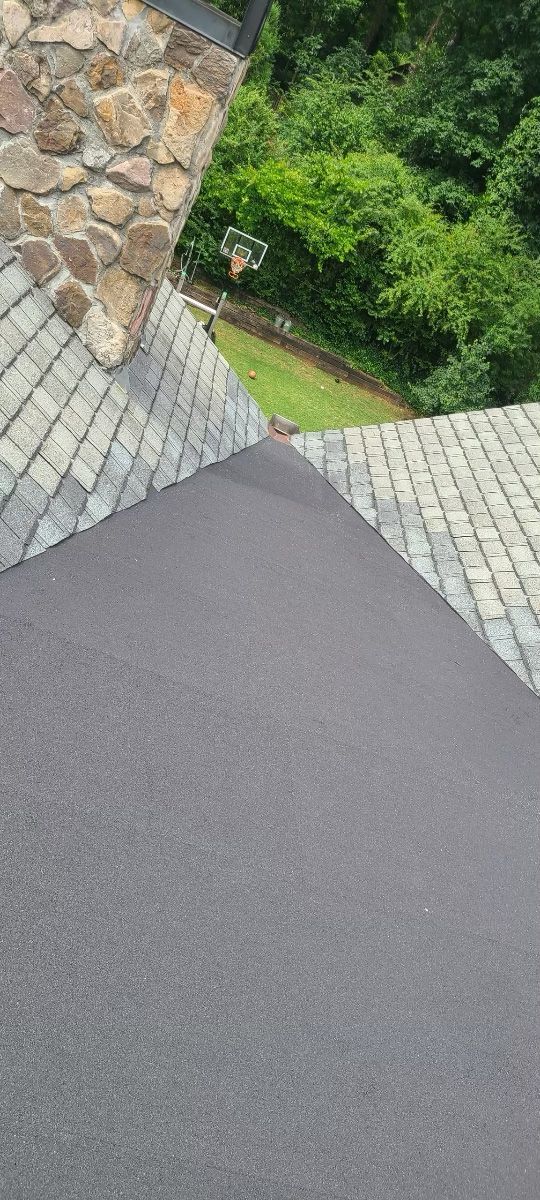 View from rooftop with dark grey asphalt shingles and light grey stone tiles, overlooking green trees and a lawn.