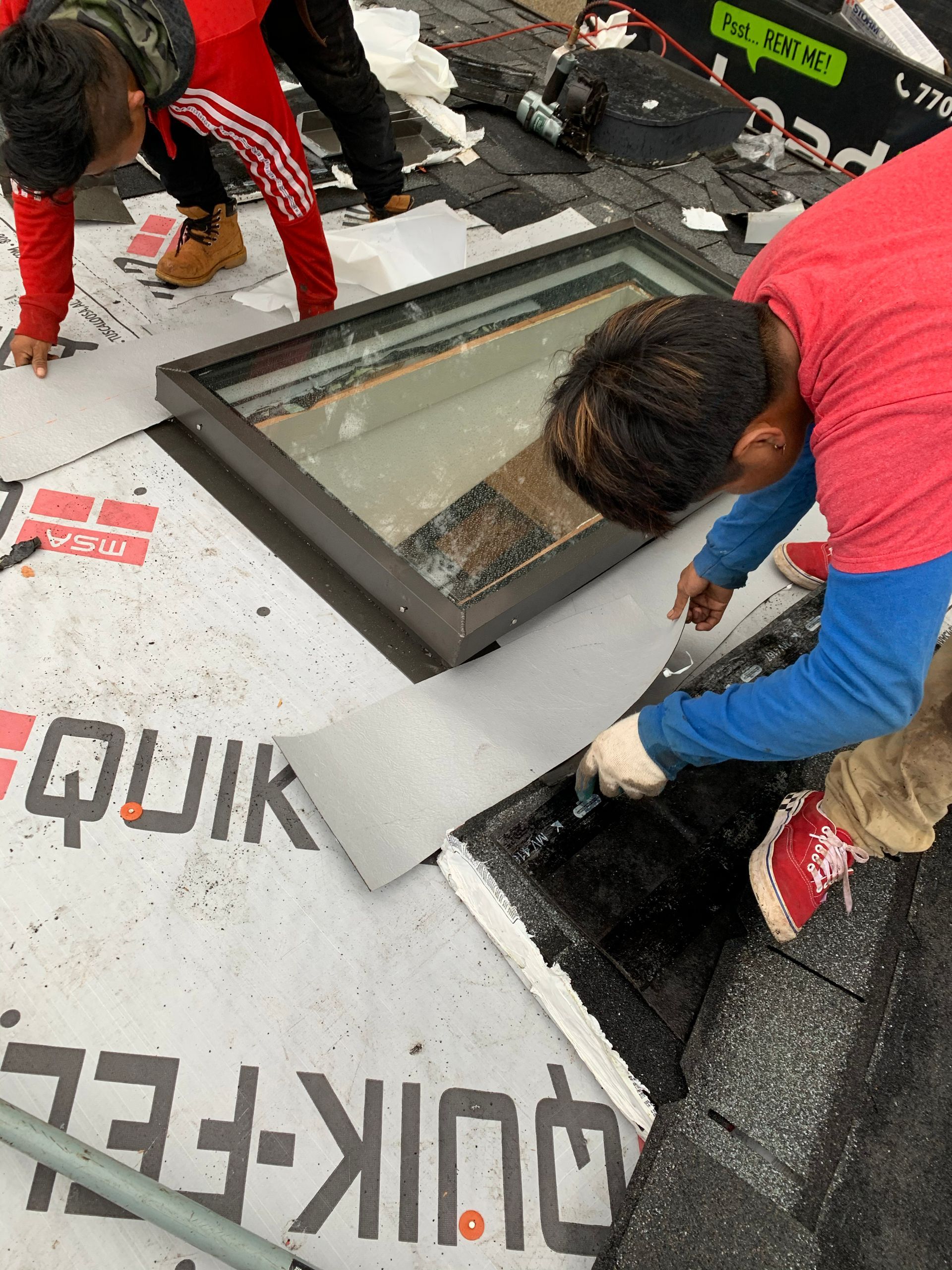 Roofers installing flashing around a skylight. One uses a knife, another lays roofing material.