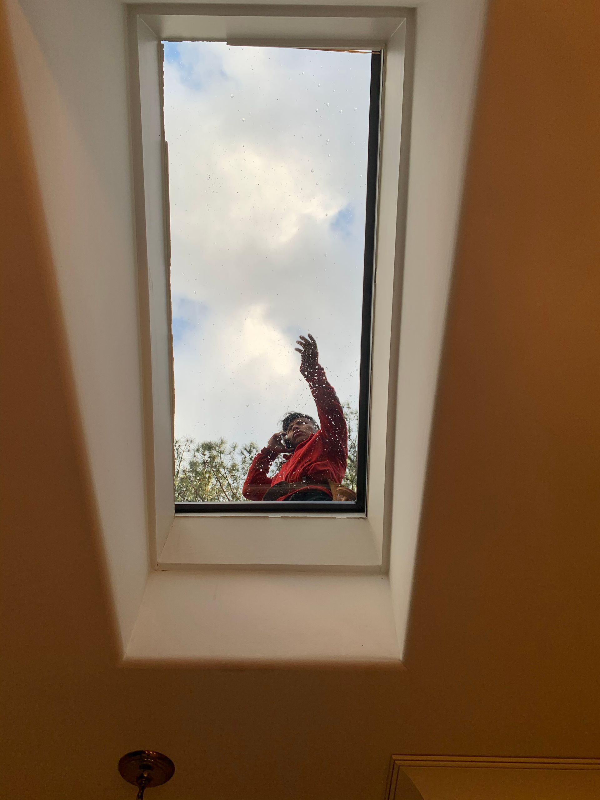Person in red shirt reaching toward a skylight, blue and cloudy sky visible.