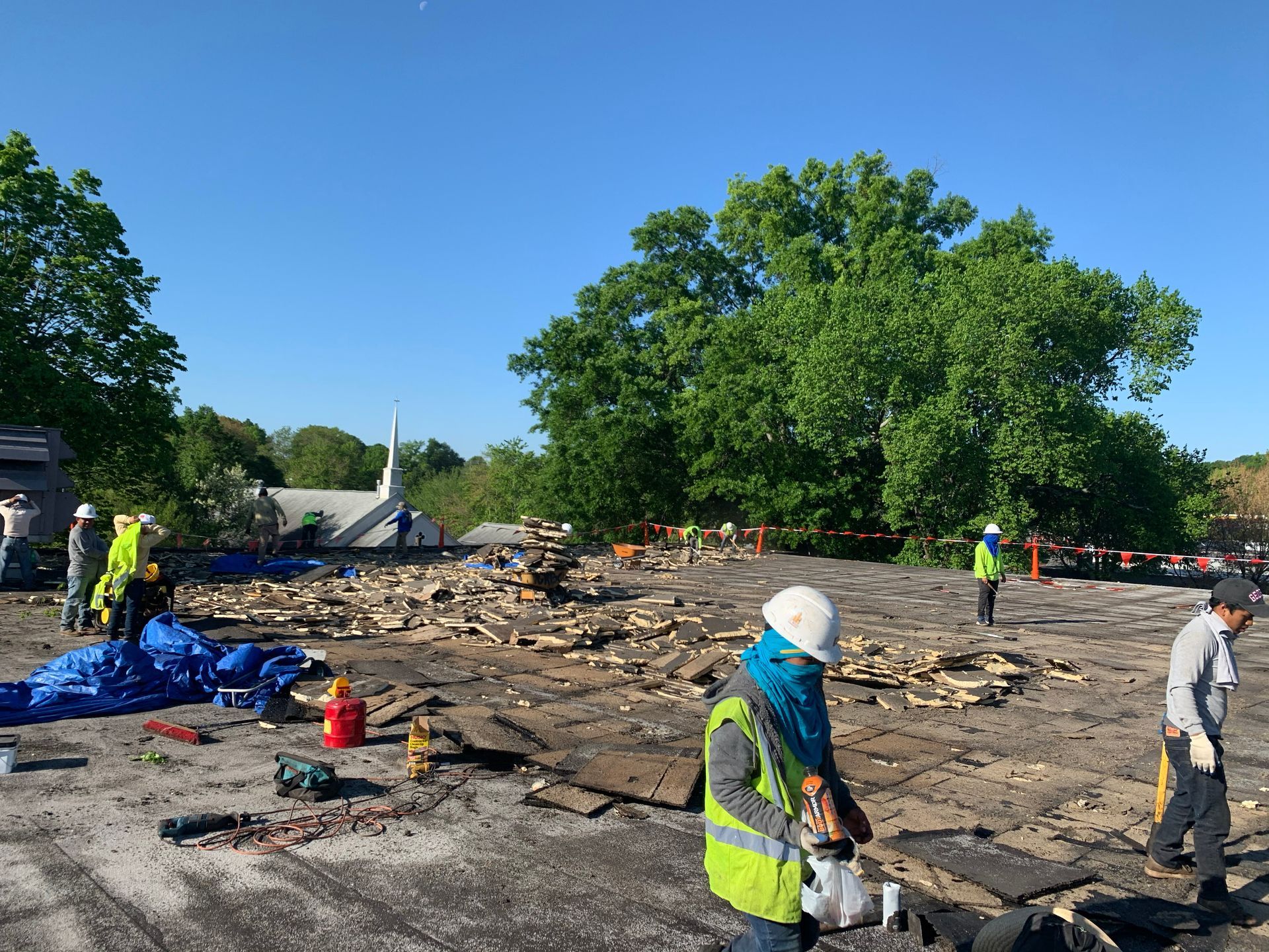 Construction workers on a roof, removing debris. Blue tarps and tools visible. Sunny day.