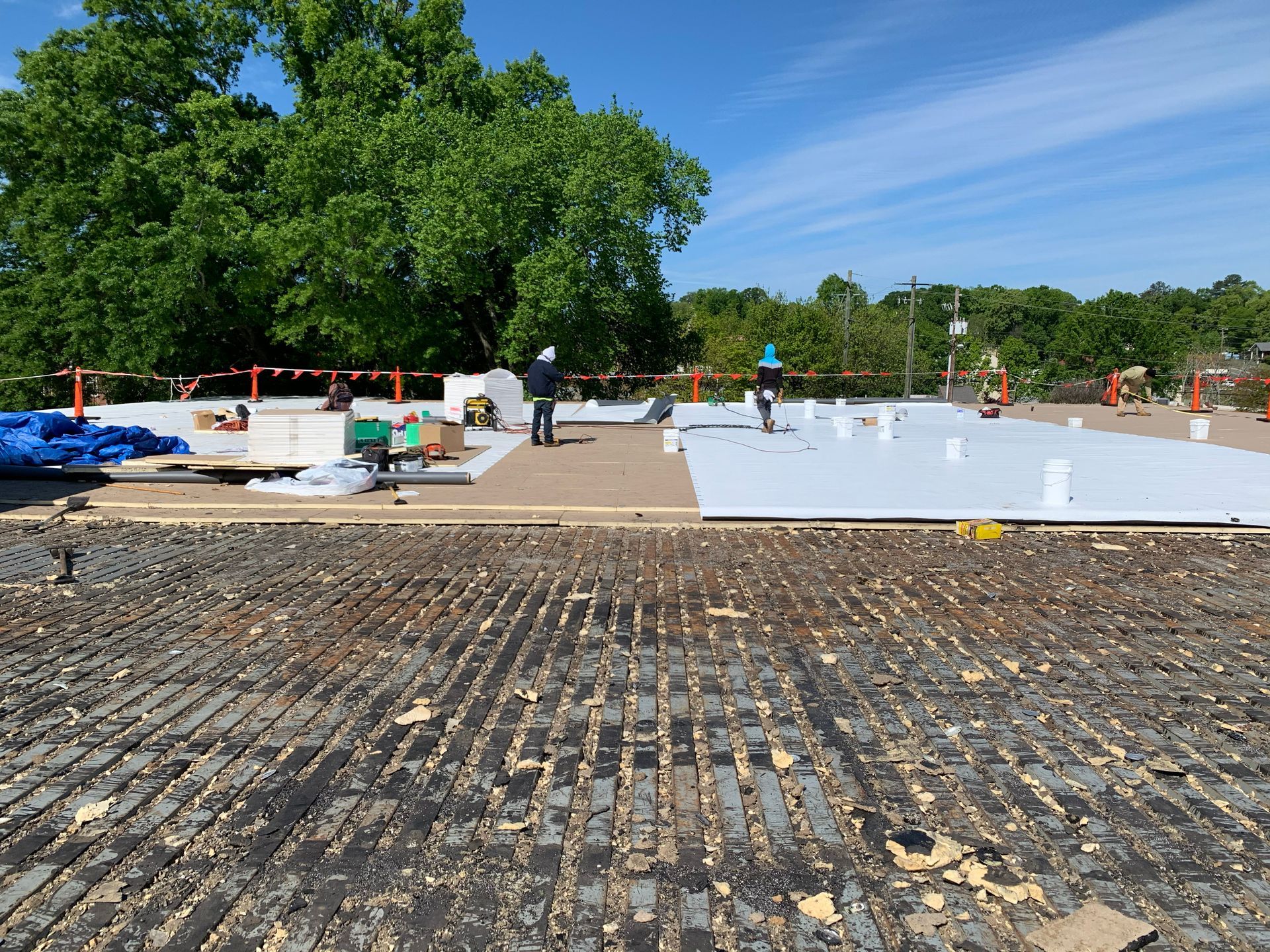 Roof construction: Workers install white panels on a flat roof. Blue tarp, green trees, and blue sky in background.