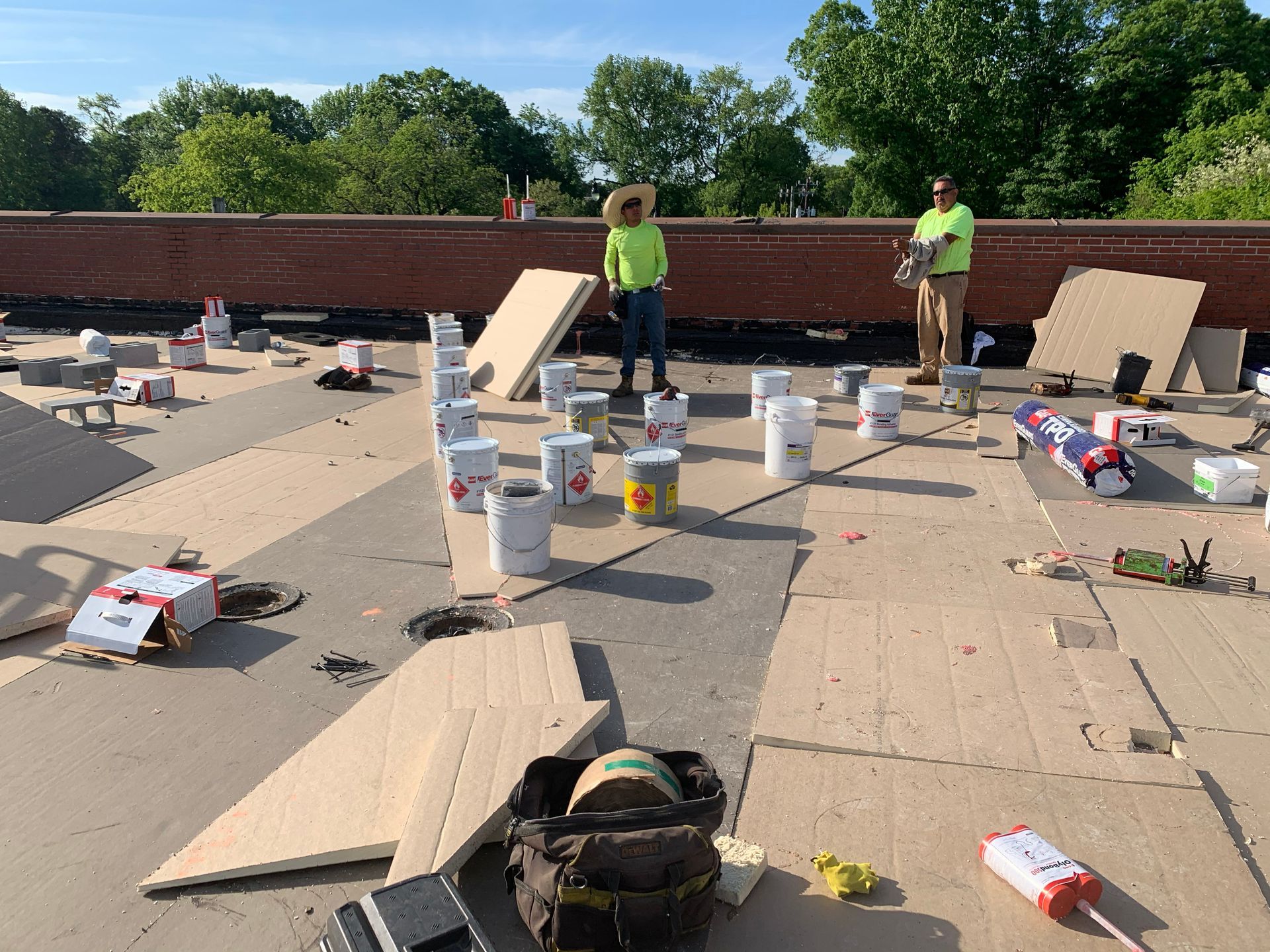 Two workers on a flat roof with materials, including paint buckets and boards. Sunny day, trees in background.