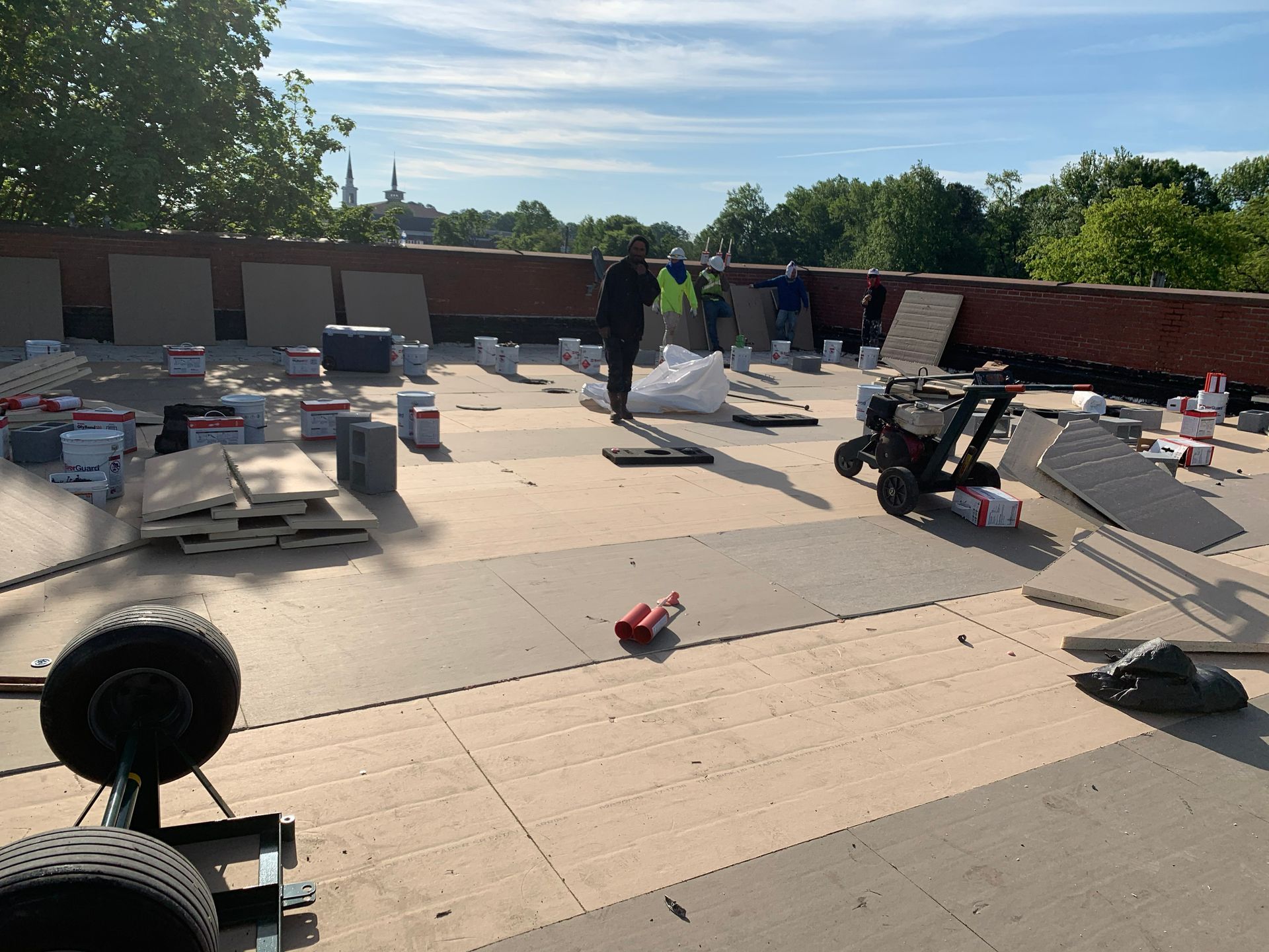 Workers installing tiles on a flat roof under a blue sky. Construction materials and equipment are scattered around.
