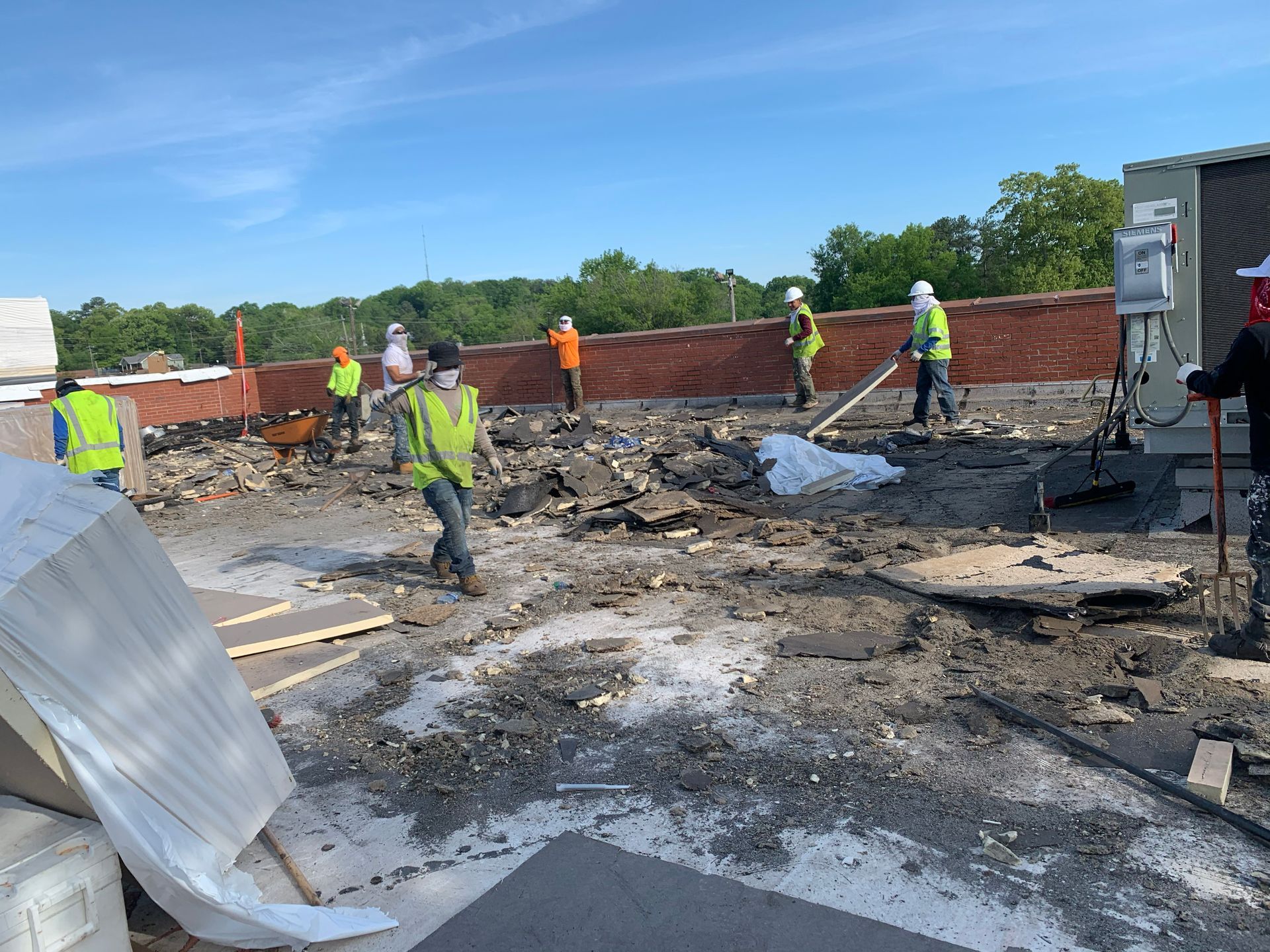 Construction workers on a roof, removing old roofing material. Workers wear safety vests and hard hats.