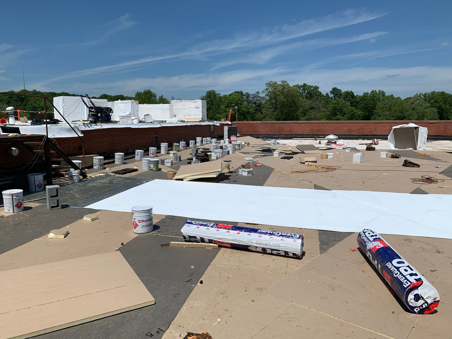 Roof under construction on a sunny day. White roofing material, tools, and supplies are scattered across the surface.