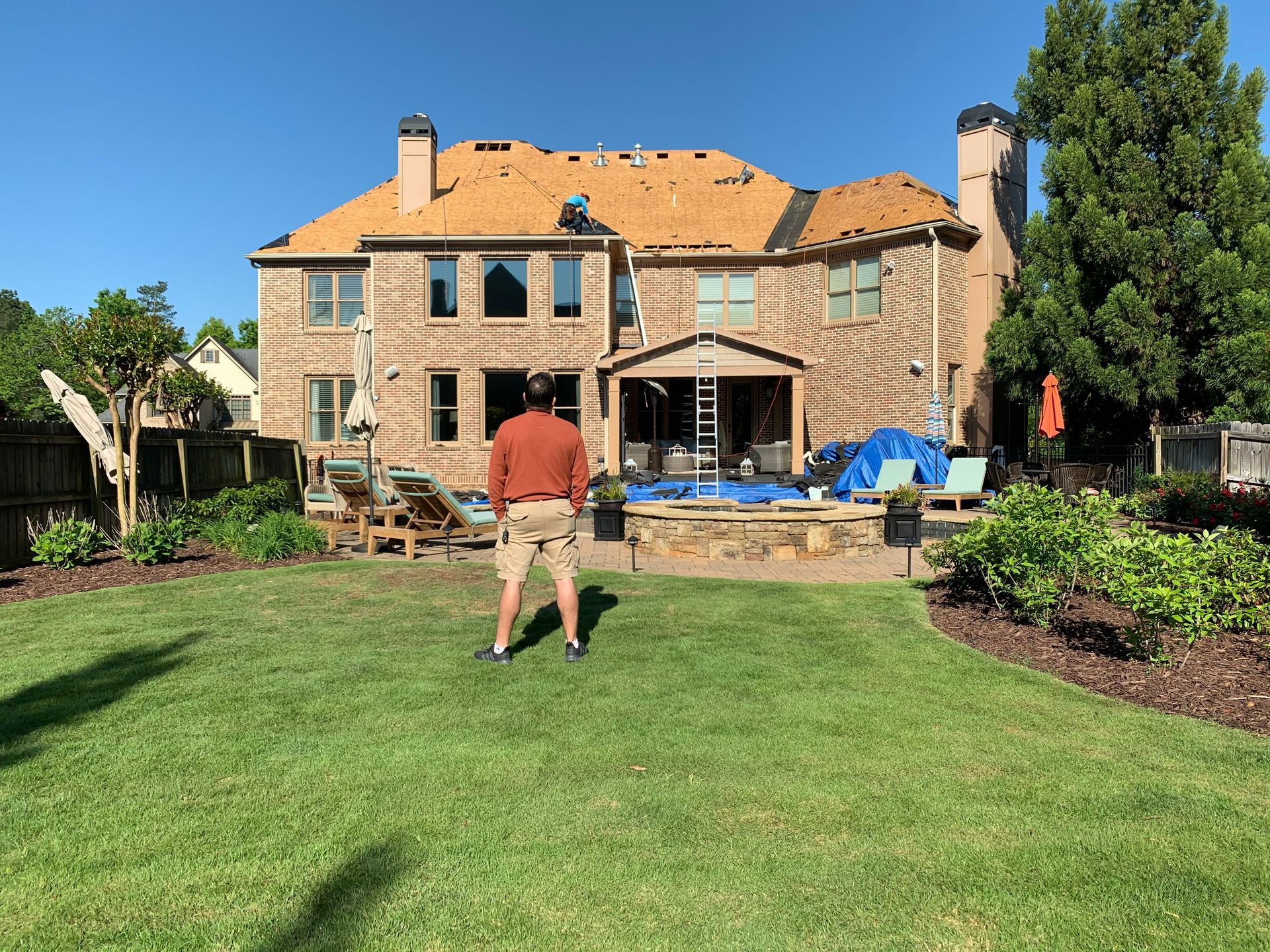 Man in backyard facing house with exposed roof; fire pit and furniture visible.