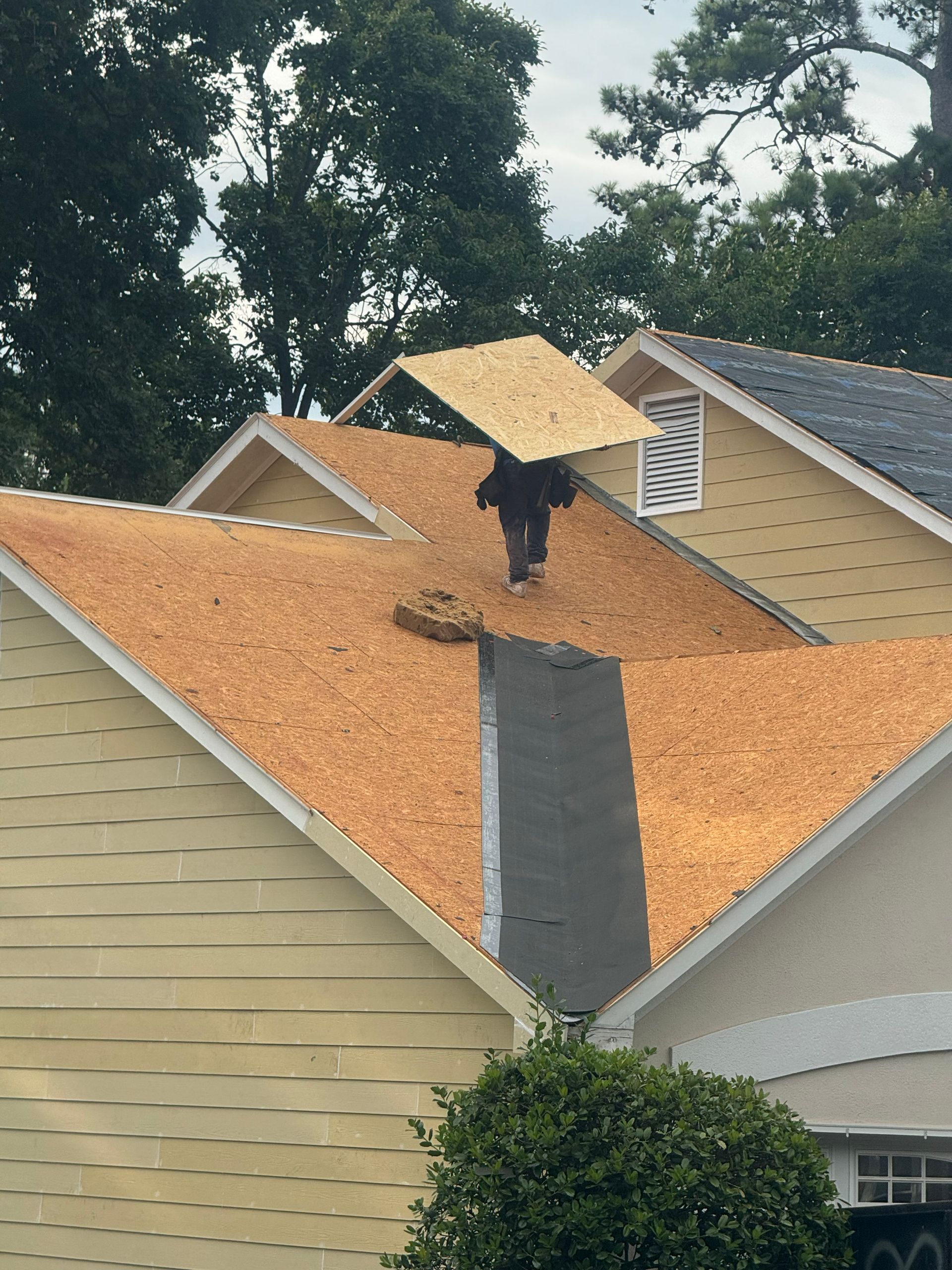 Roofer carrying a large piece of roofing material on a house roof. Beige, brown, and gray colors are visible.