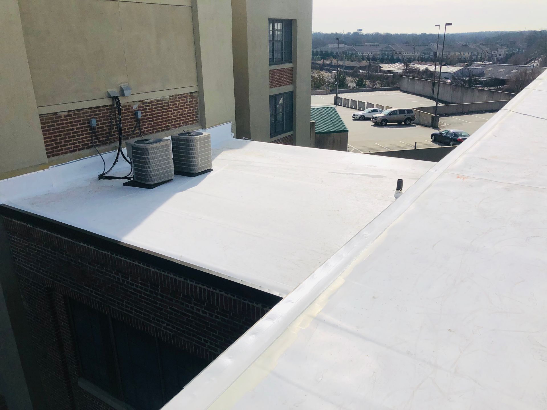 Flat white roof with HVAC units, adjacent to brick and beige building. Cars and distant buildings visible.