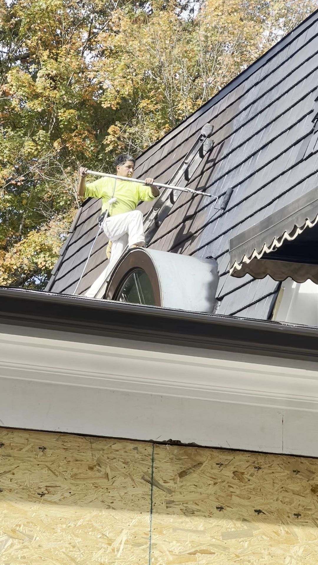 Person on a ladder painting a roof with a roller. The person wears a neon vest.