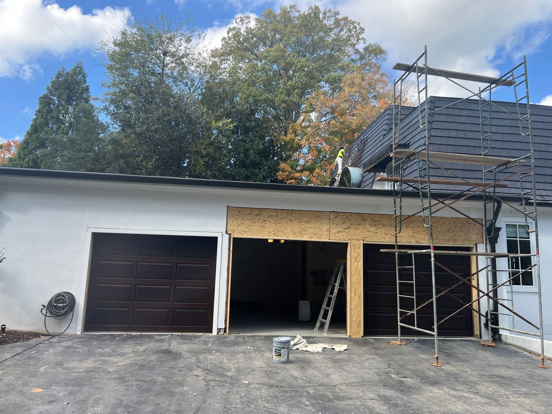 Garage exterior during renovation; two garage doors, one removed. Scaffolding, trees, blue sky.
