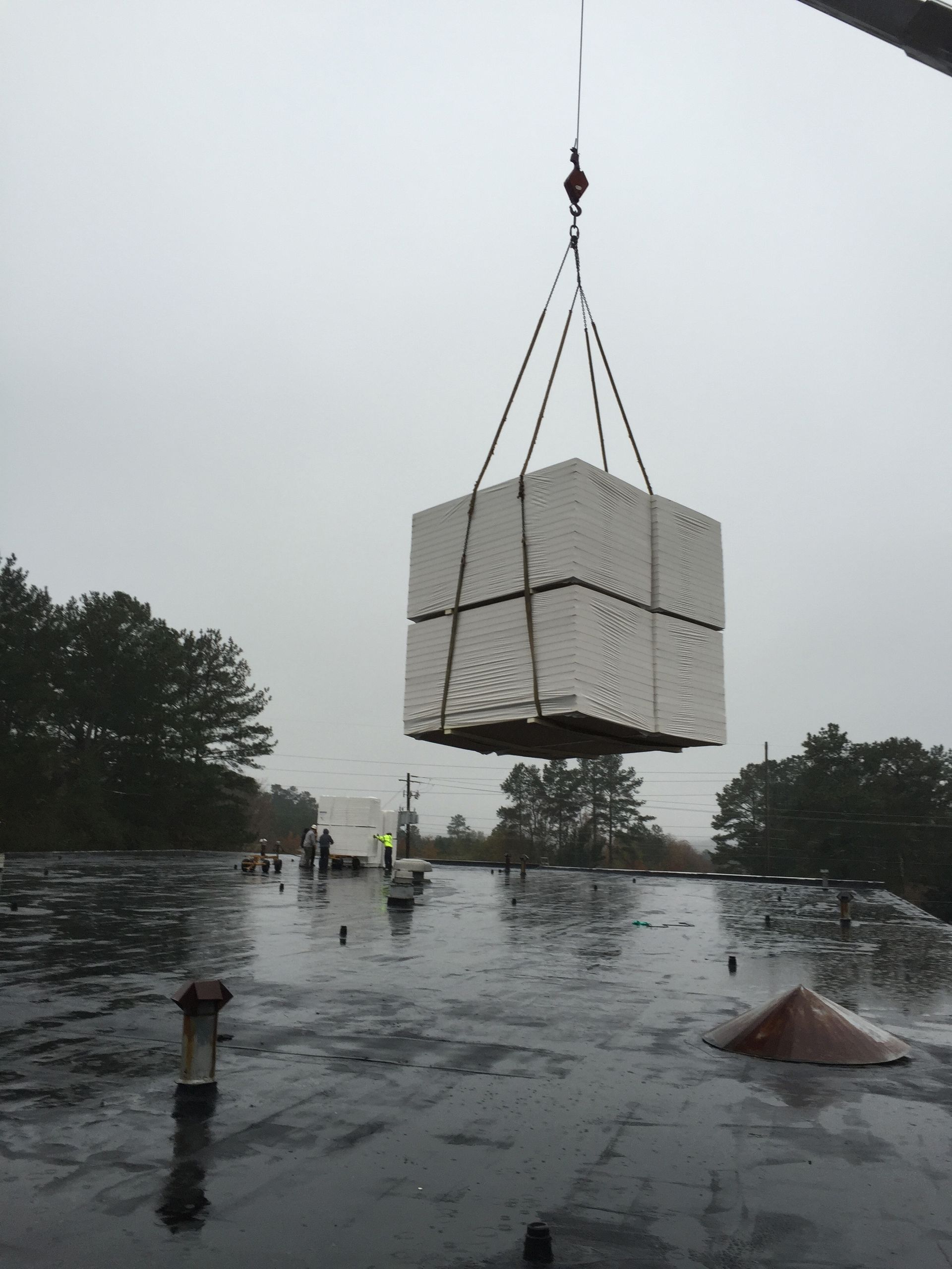 Crane lifting a stack of white insulation panels onto a wet, flat roof on an overcast day.