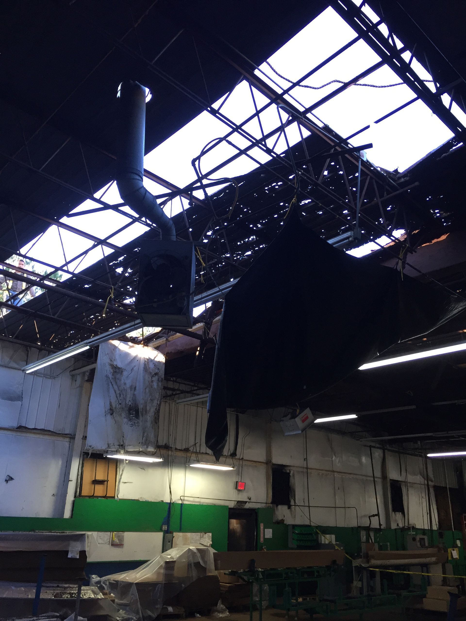 Damaged warehouse interior. Roof partially collapsed, debris scattered, exposed wires. A large vent hangs from ceiling.