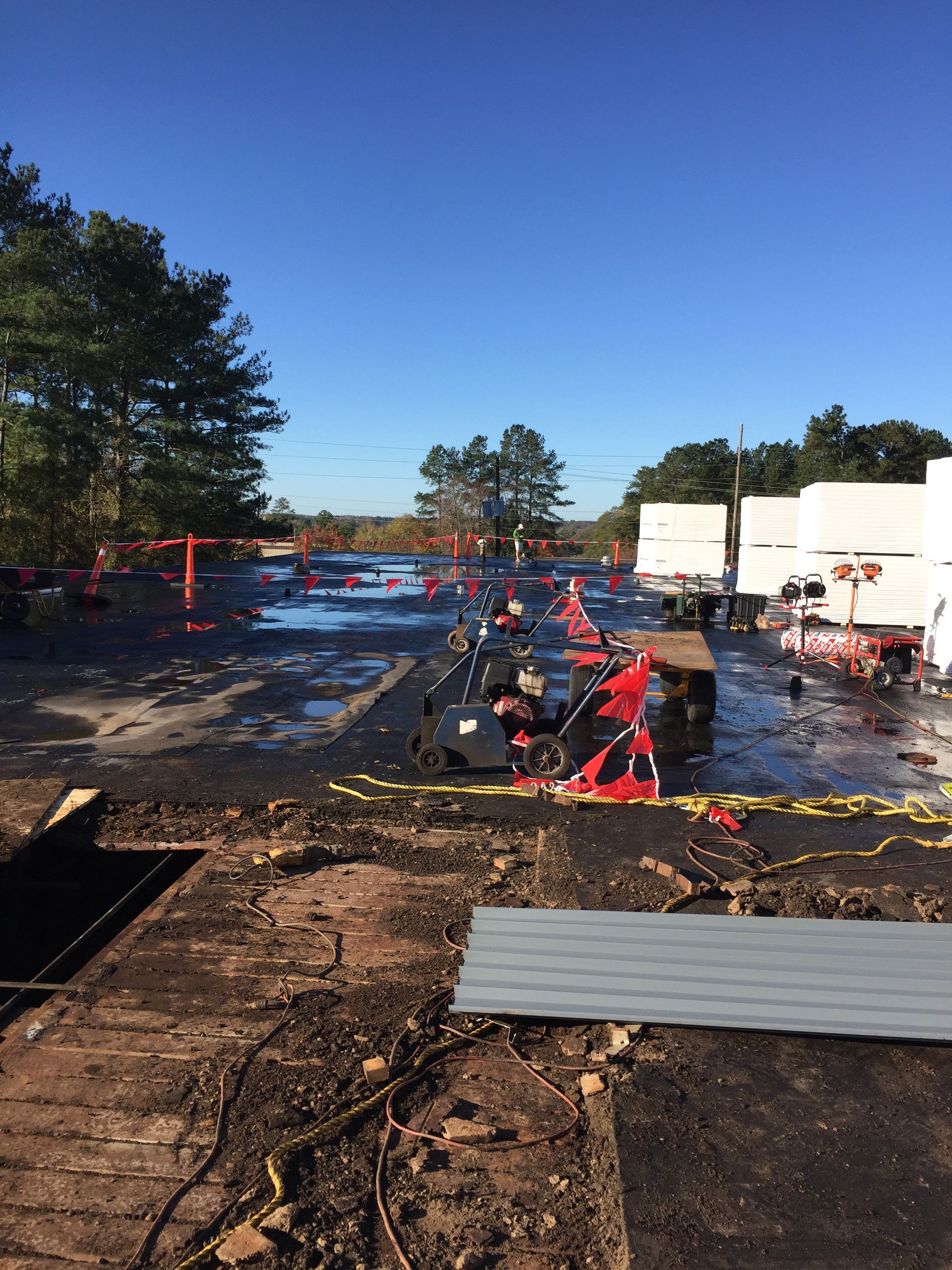 Debris-covered parking area with orange cones, possibly post-fire. White containers and trees in background under a blue sky.