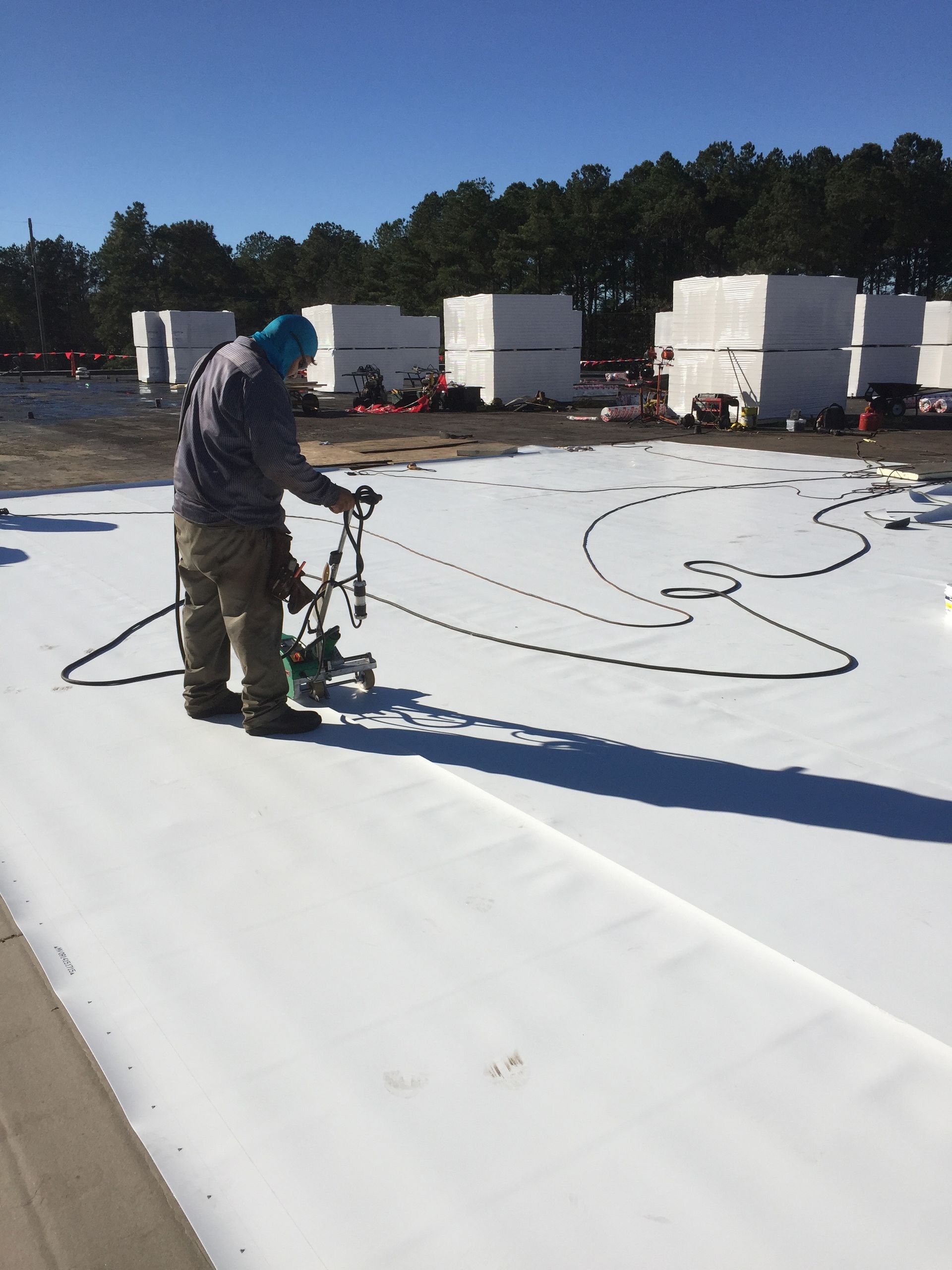 Person welding white material on a construction site; multiple rolled materials and equipment visible.