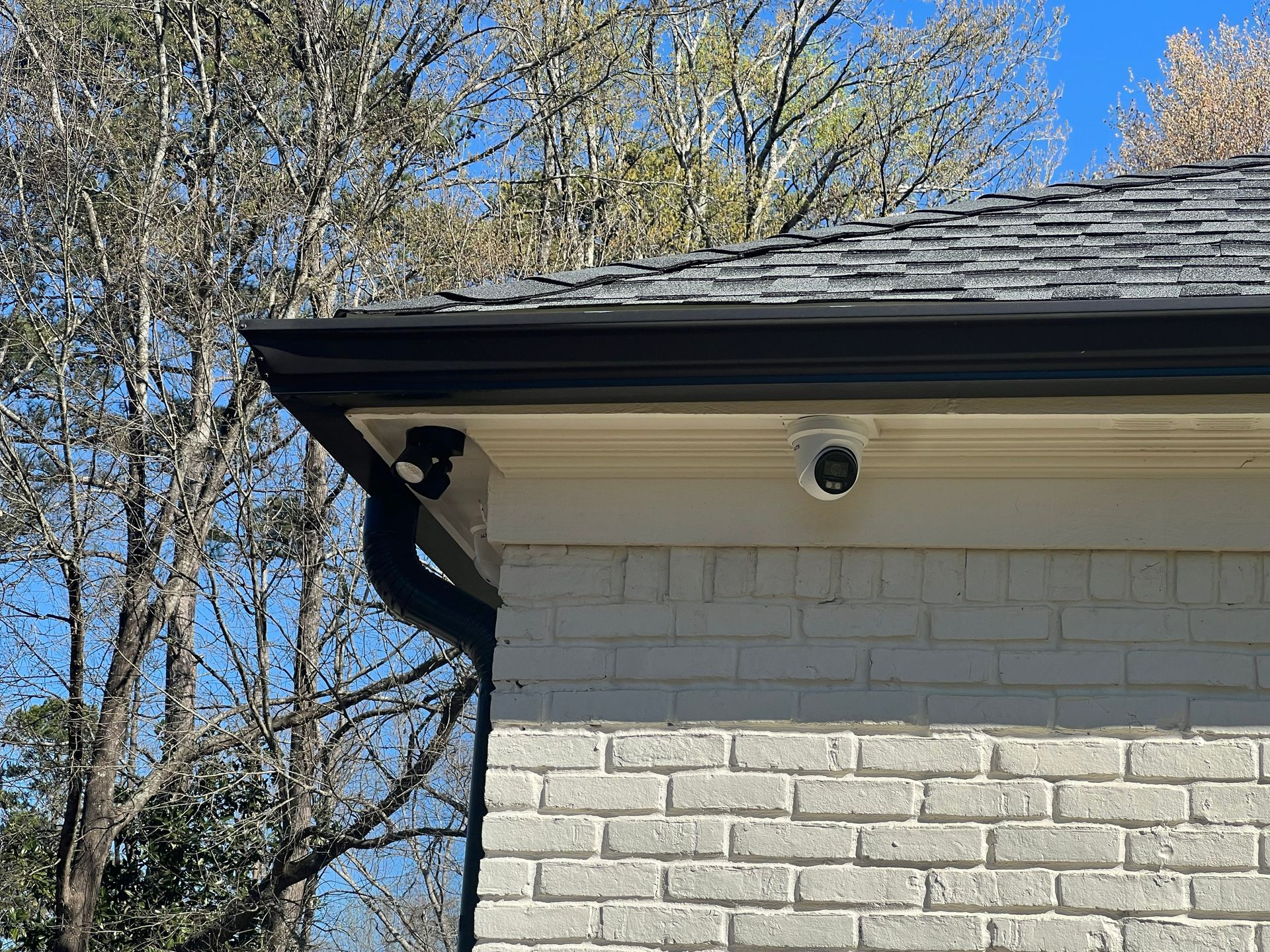 Security cameras mounted on a brick house under the roof's edge, with a black gutter and blue sky backdrop.