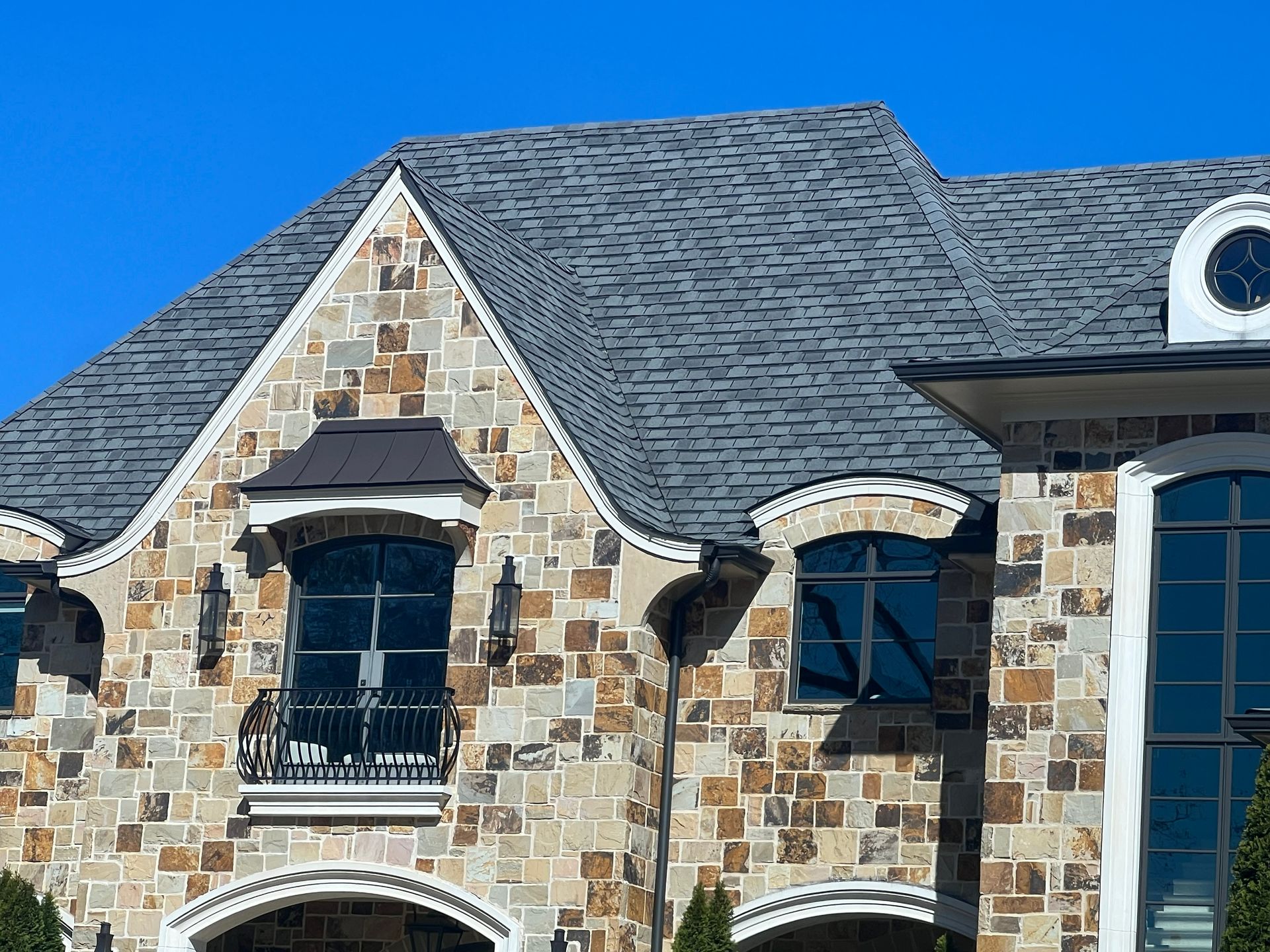 Stone-covered house with a gray shingle roof, arched windows, and a clear blue sky.