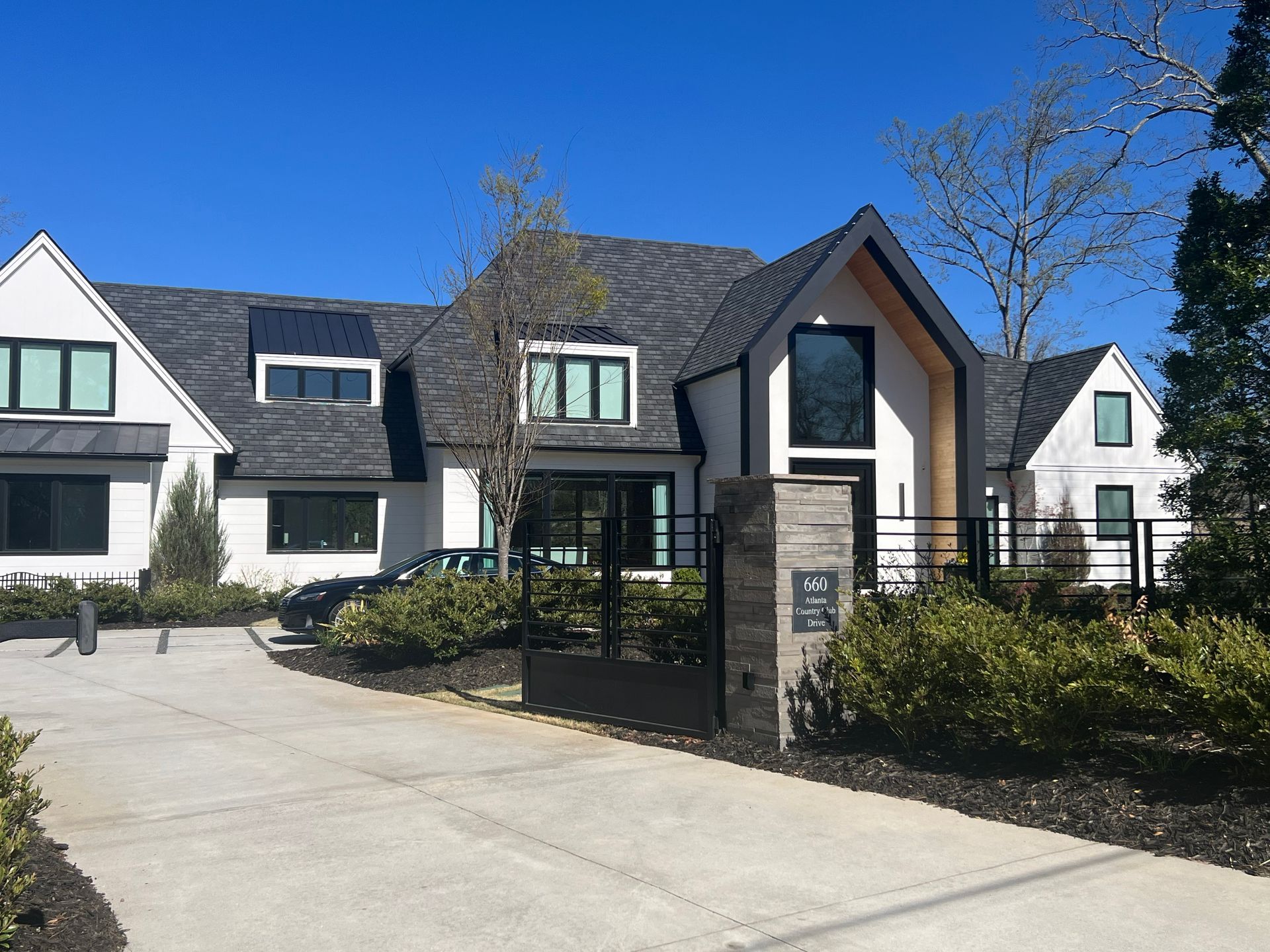 Modern white house with black roof, windows, and gate. Driveway and landscaping. Sunny day.