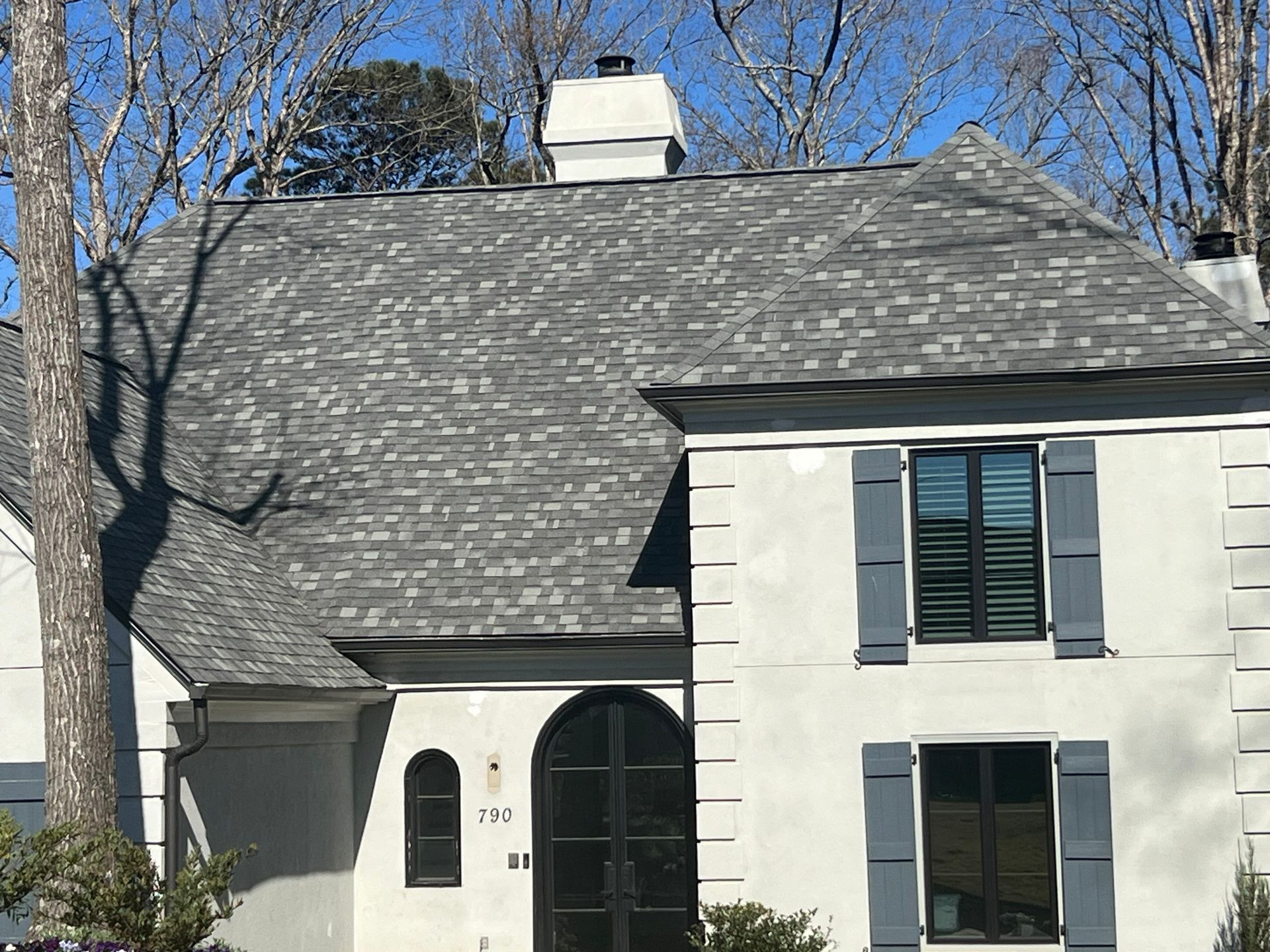Gray-roofed house with shutters, stucco exterior, and black front doors. Sunny day, bare trees.