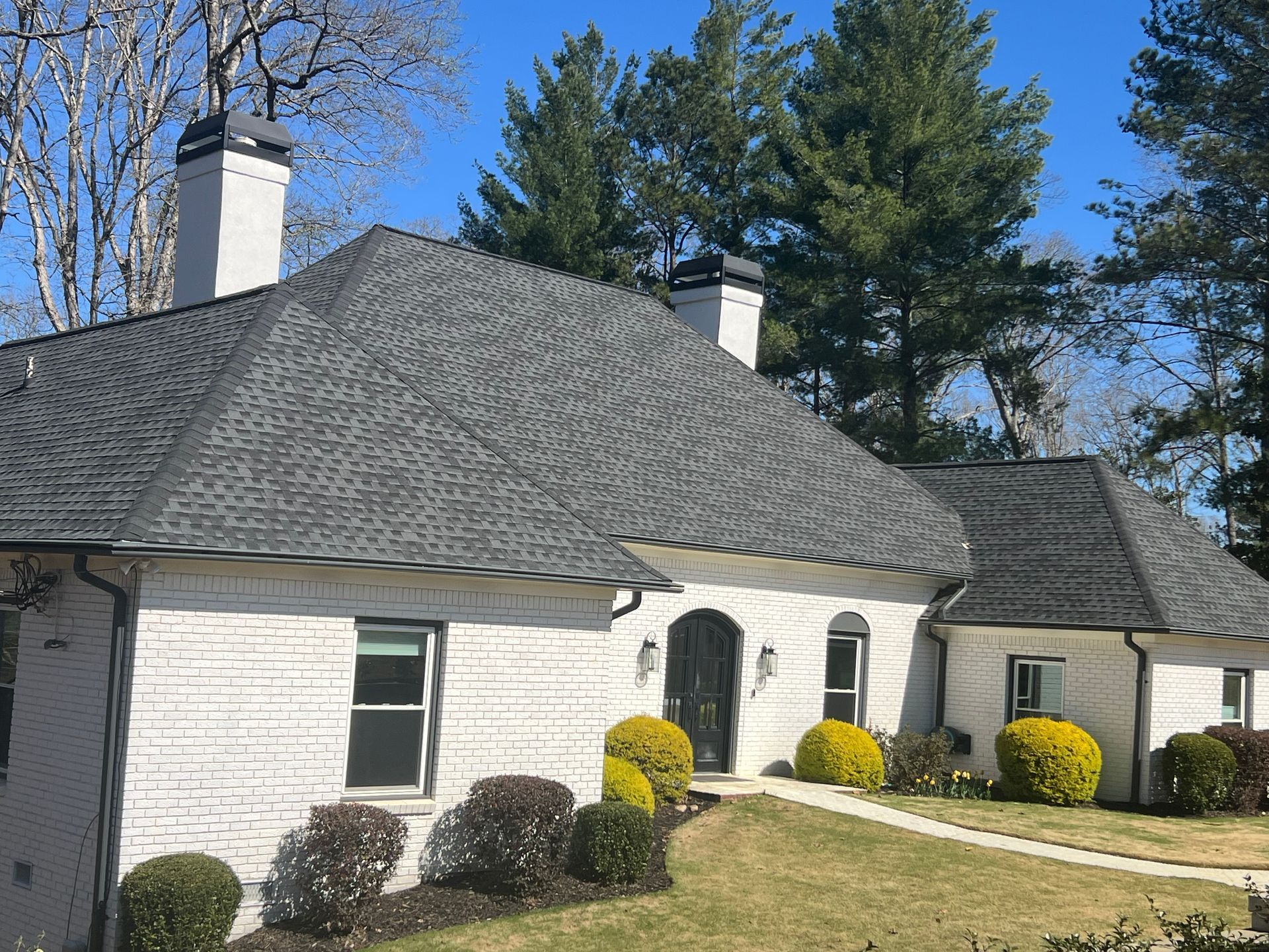 White brick house with gray roof and chimneys. Green shrubs line the walkway.