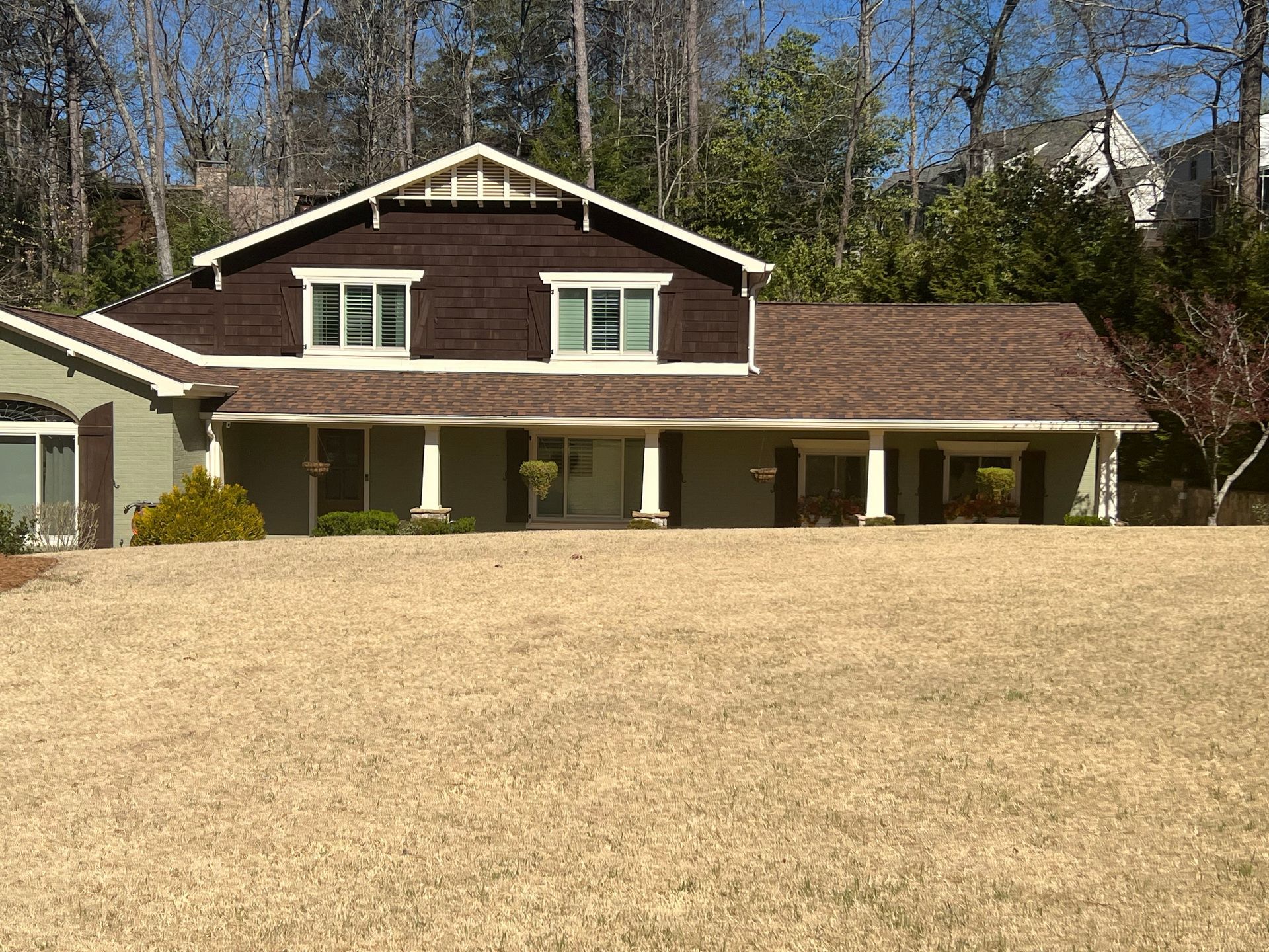 Two-story brown house with white trim and a long porch, set in front of a dry, brown lawn and trees.