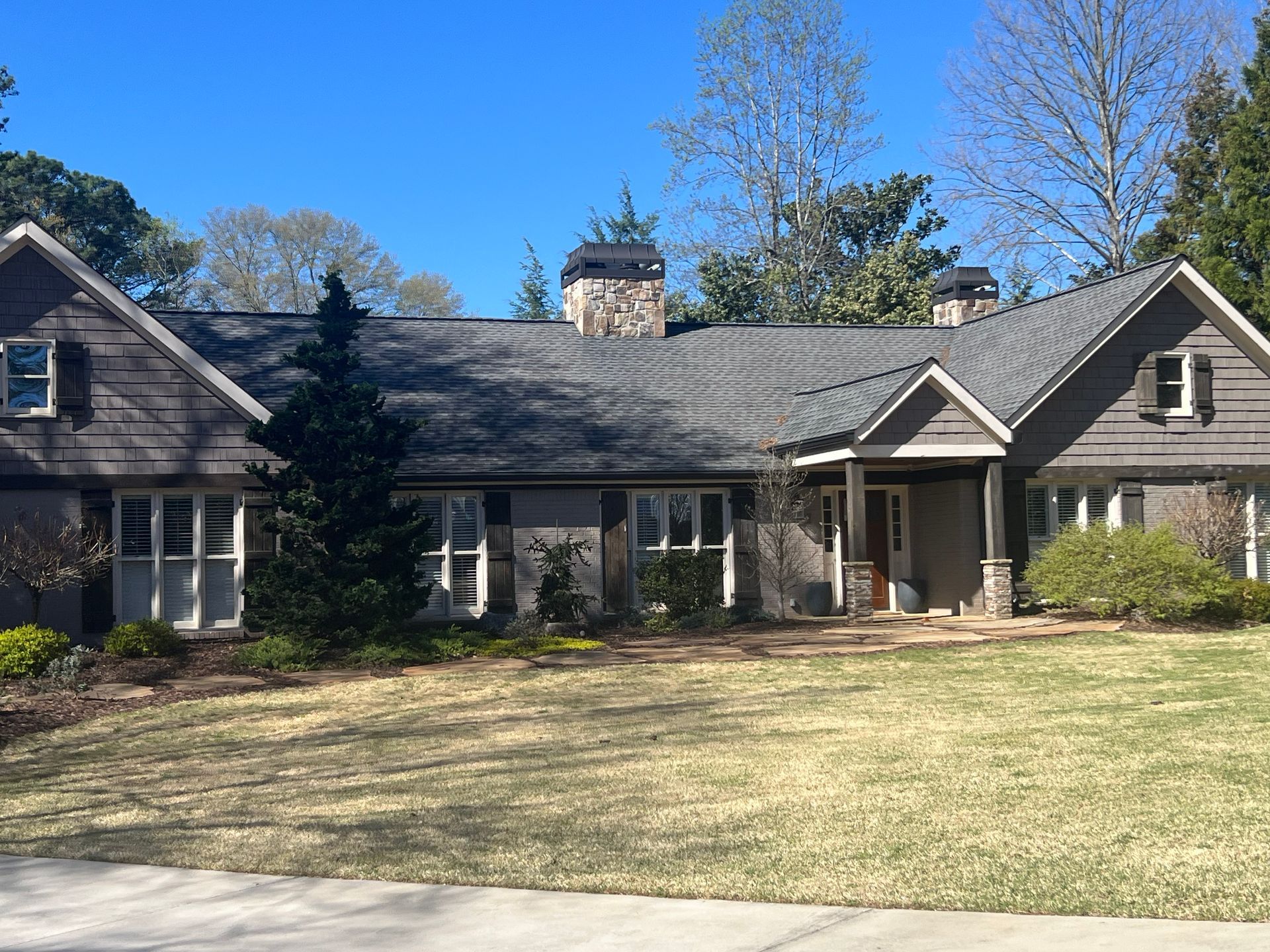 Brown-shingled house with multiple rooflines, stone chimney, and shutters, set on a lawn with trees under a blue sky.