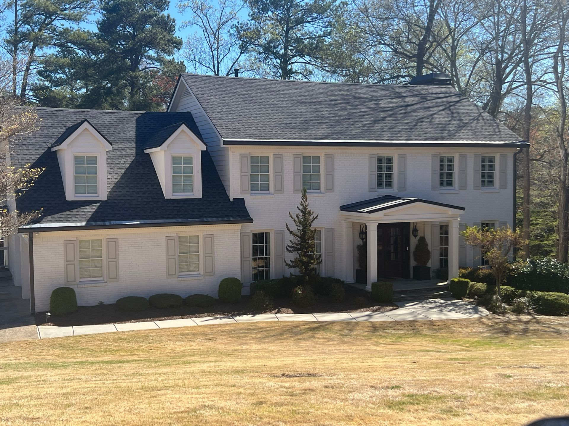 White two-story house with black roof, shutters, and porch. Dormers on the roof. Lawn in the foreground.