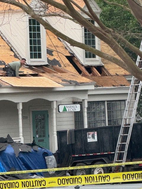 Man on a roof repairs shingles. A dumpster, ladder, and caution tape are nearby.