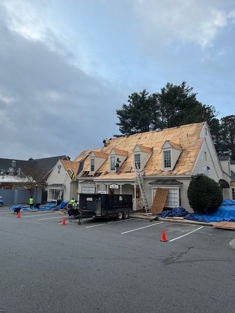 Roofers working on a building with three dormers, blue tarps on the ground, and a cloudy sky.