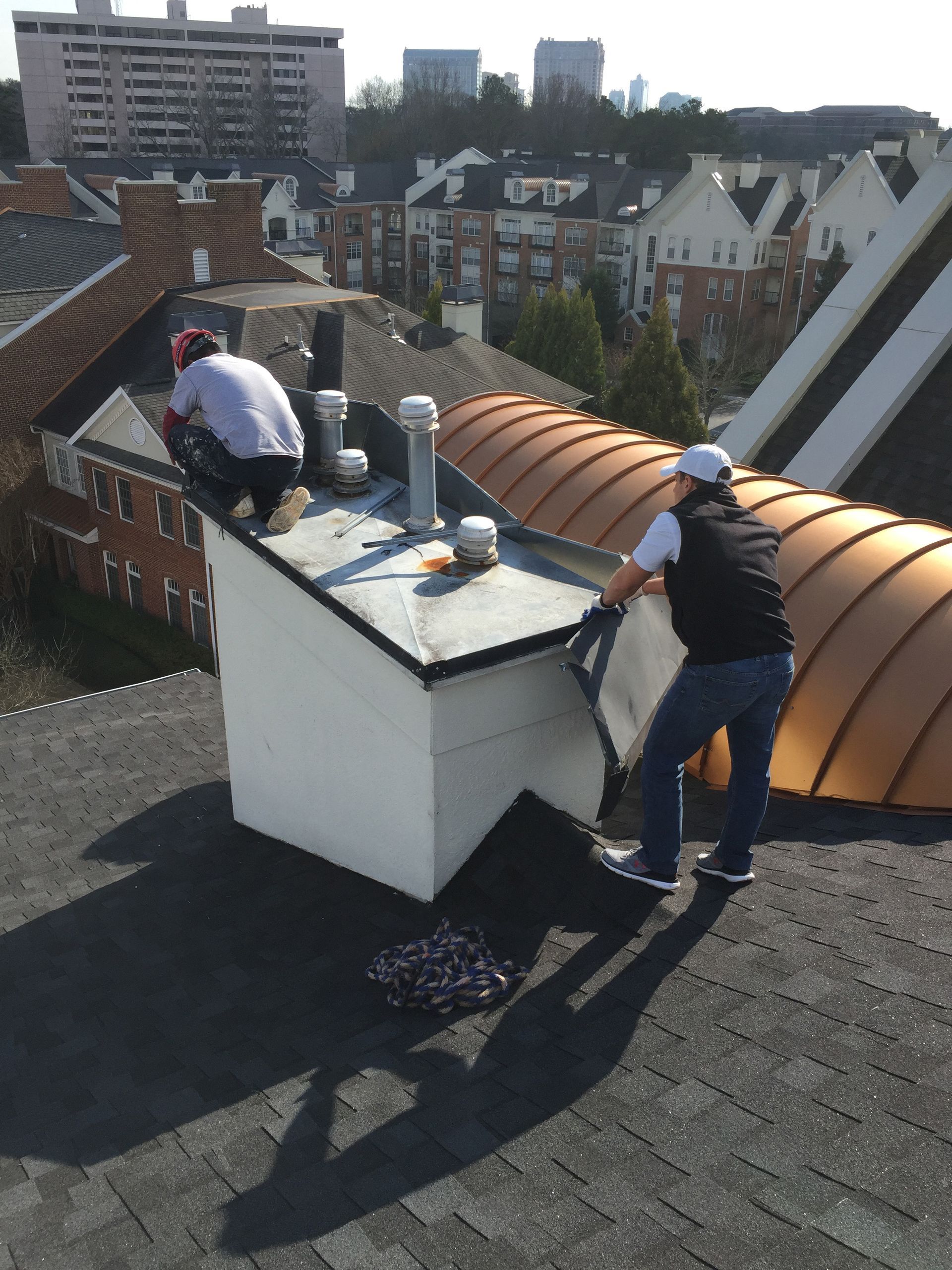 Two people working on a roof, near a chimney. One person is installing some kind of covering. Sunny day.