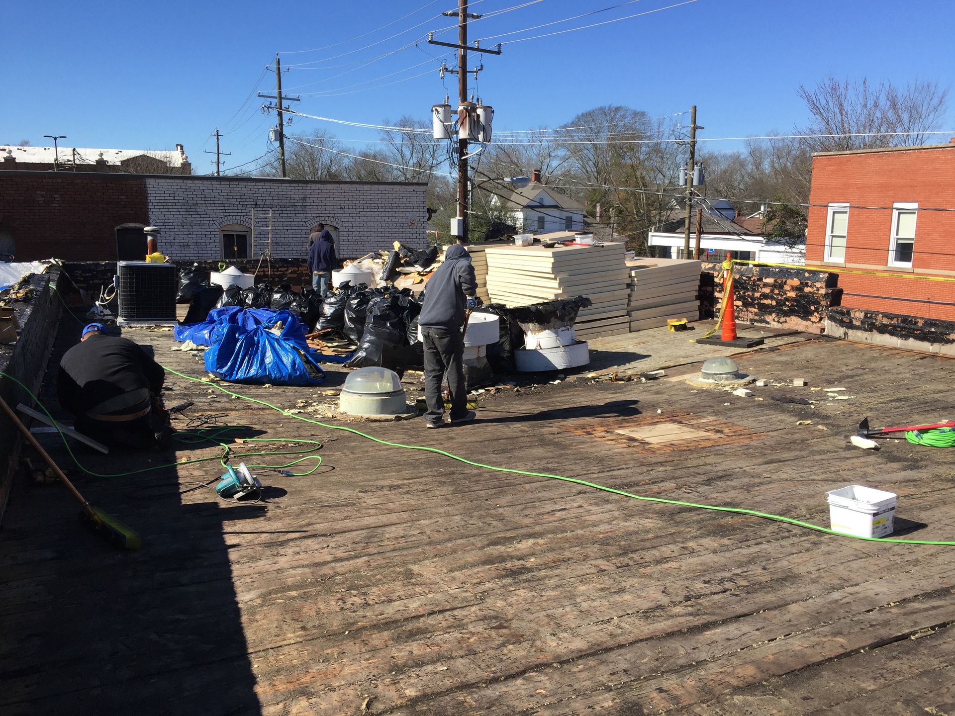Workers on a rooftop, installing corrugated metal. One uses a broom, another holds a tool, and another is on the left.