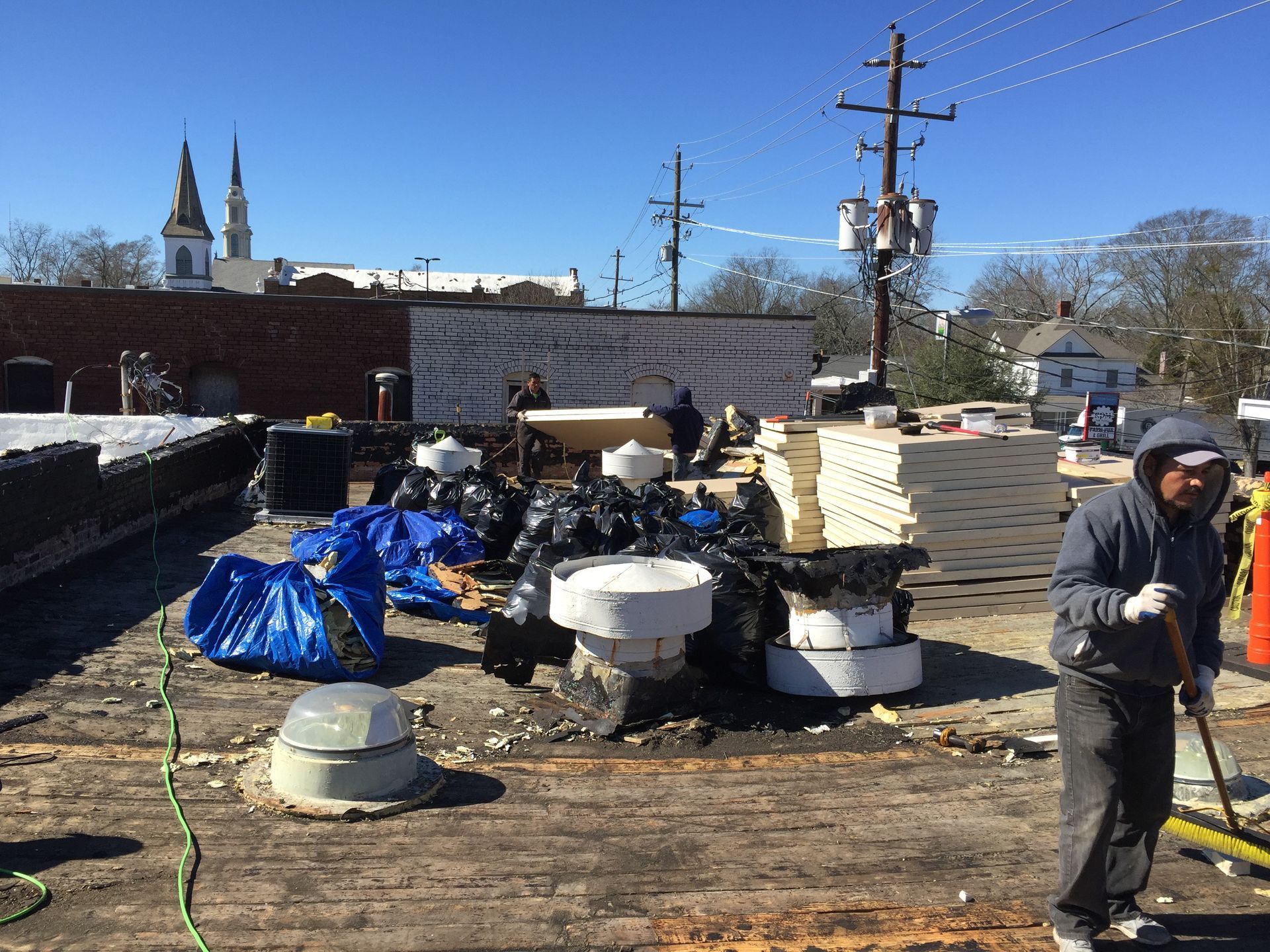 Roofing crew working on a flat roof. Several piles of debris, supplies, and a worker sweeping debris with a brush.