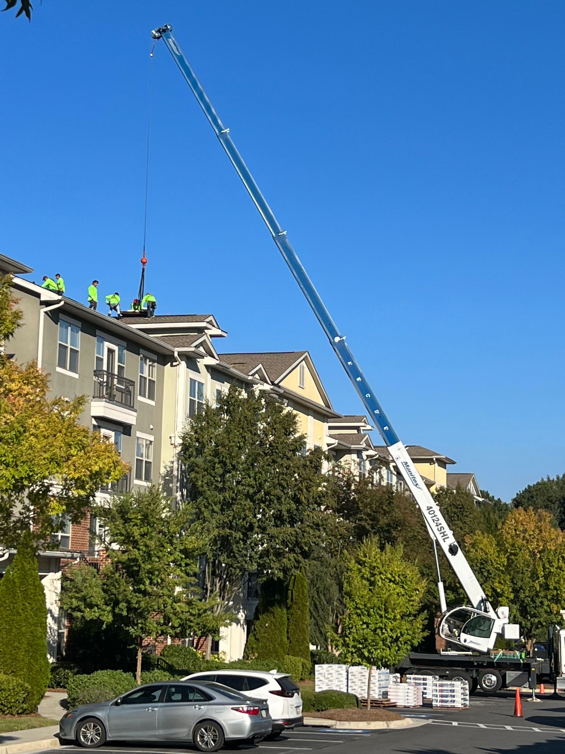 Crane lifting materials onto a building roof. Workers in green vests on rooftop under a clear blue sky.
