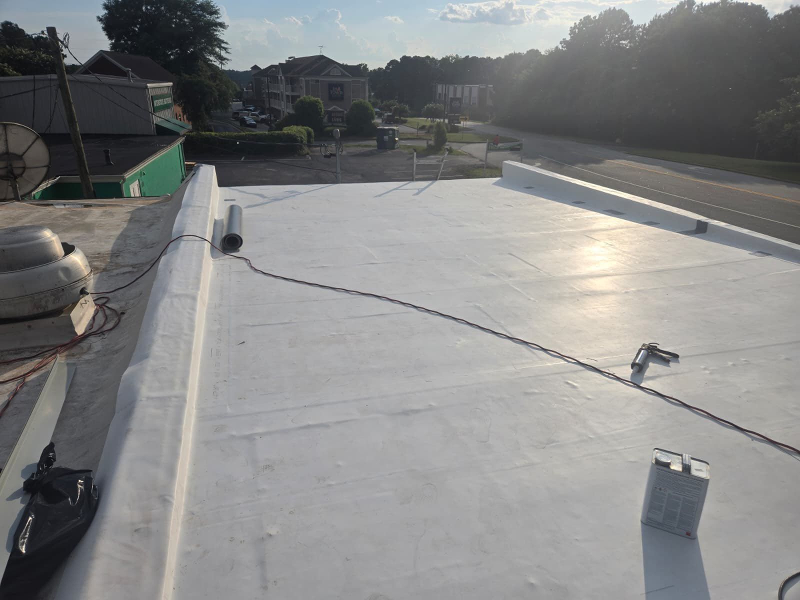Flat, white commercial roof, possibly being repaired. Road and houses visible in the background. Sunny.