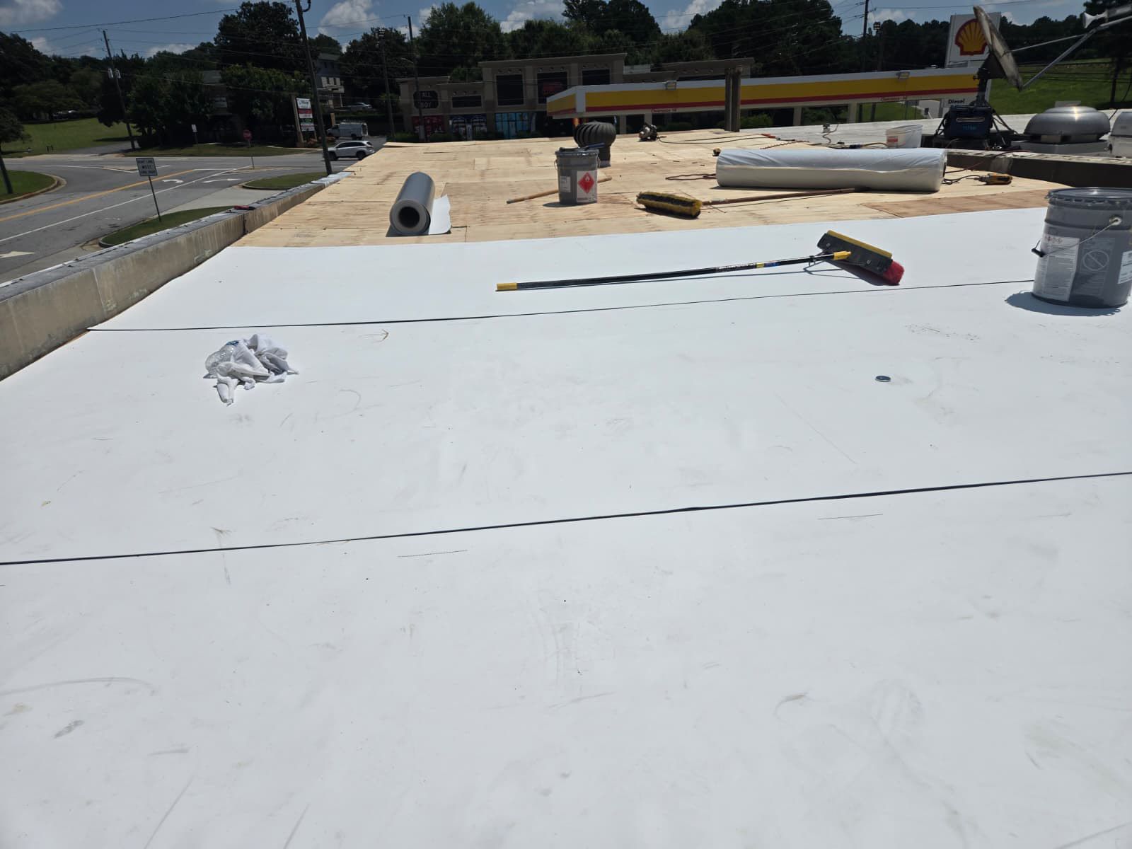 Flat roof construction site with white membrane being installed, tools, and gas station in the background.