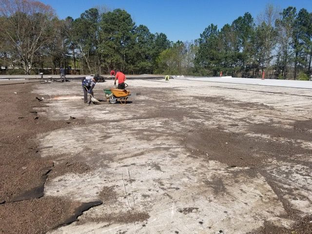 Workers spread gravel on a large, flat surface. Trees and blue sky in background.