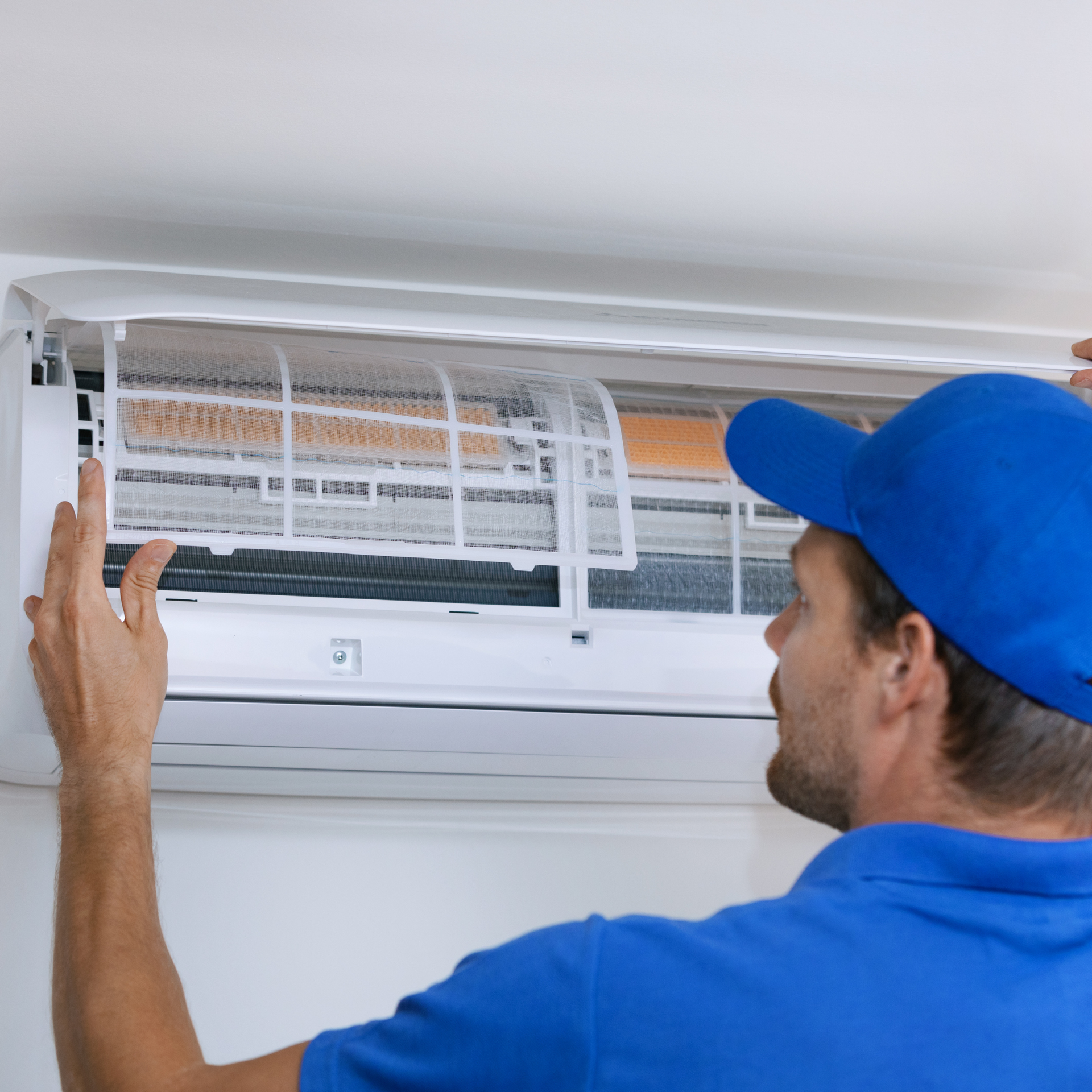 Man in blue uniform examining an air conditioning unit filter. Indoors, white setting.