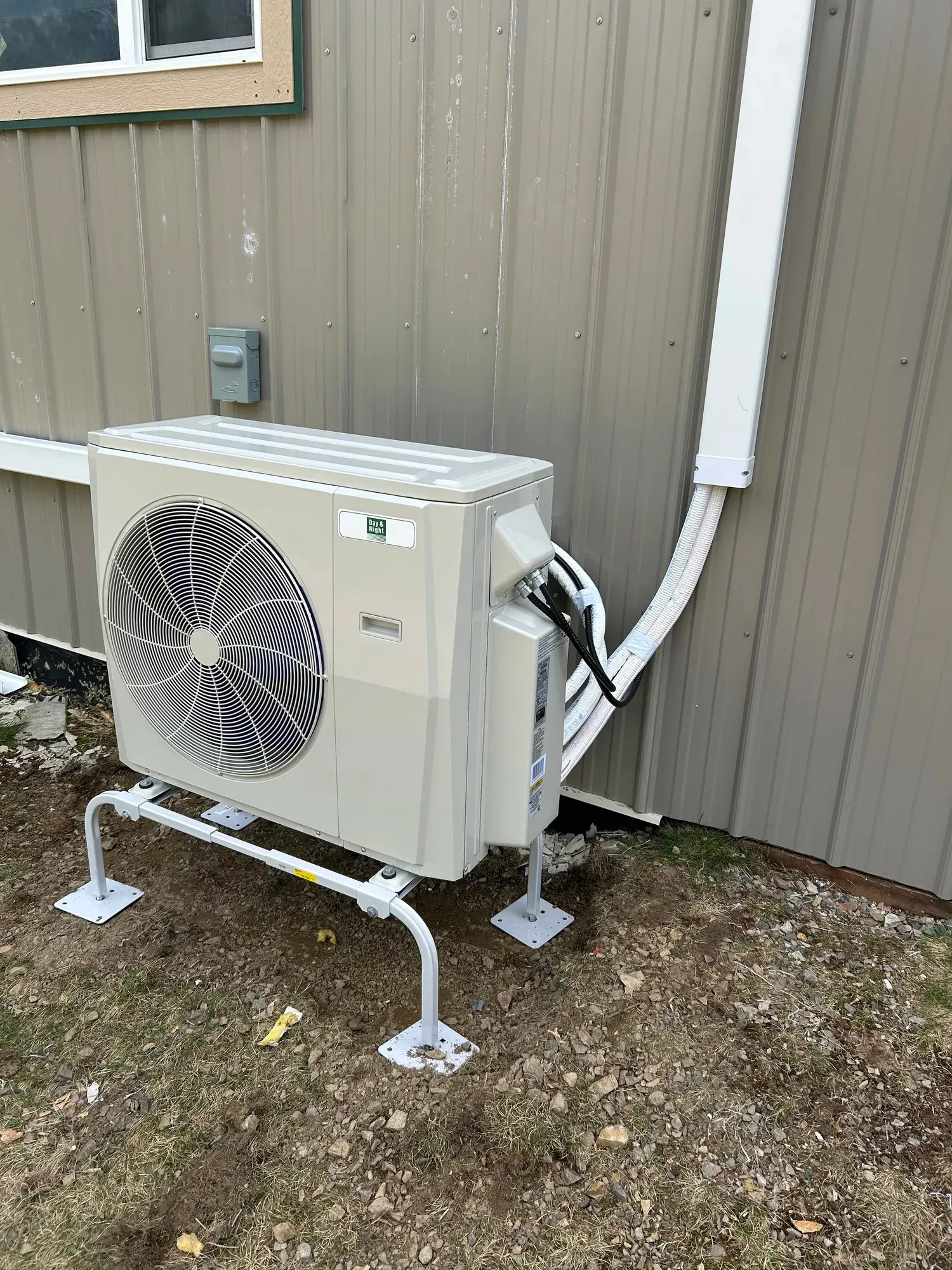 Heat pump unit mounted on metal legs beside a building with beige siding and a white conduit.