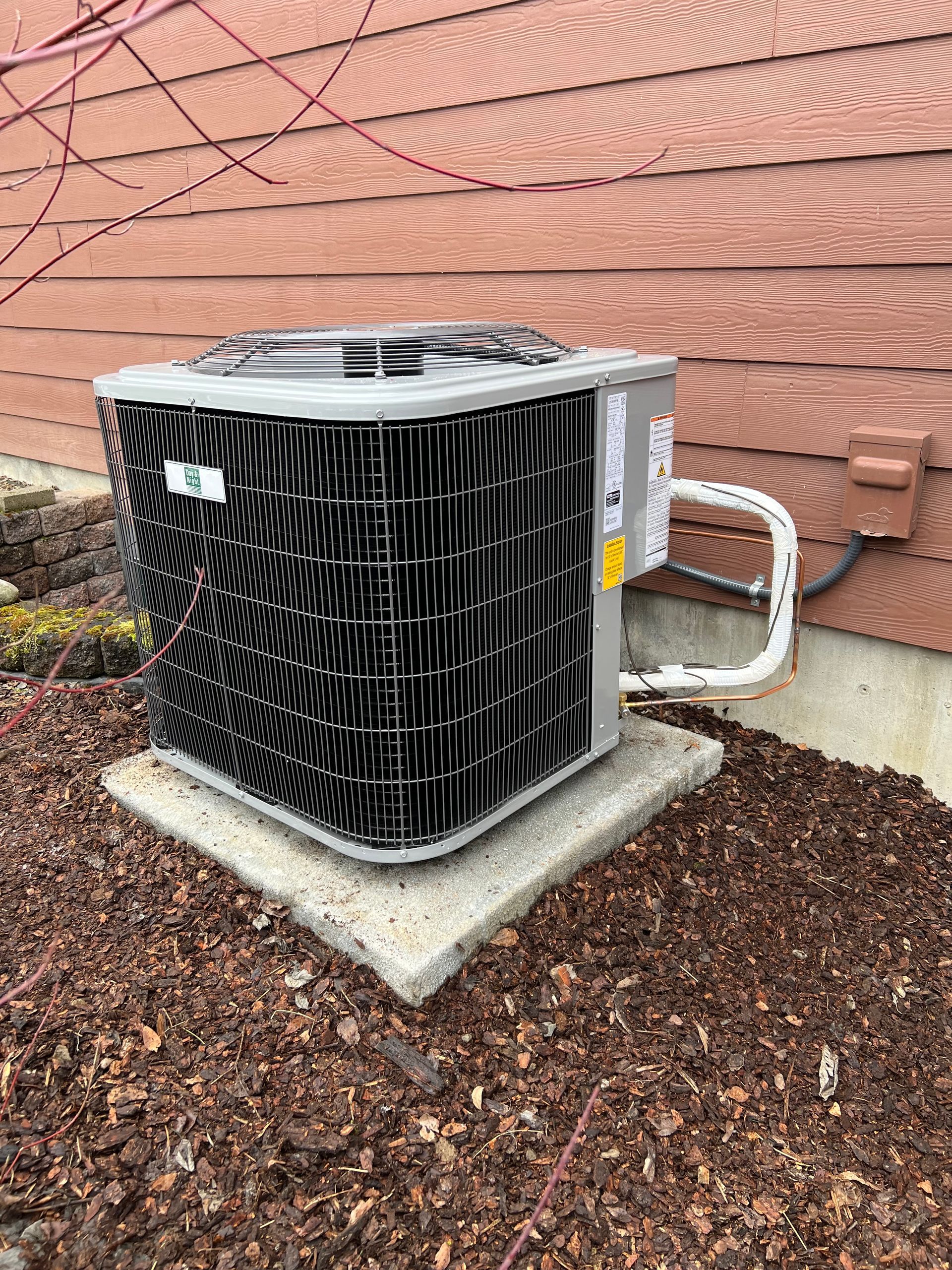 Air conditioning unit on a concrete pad, brown siding backdrop, mulched ground.