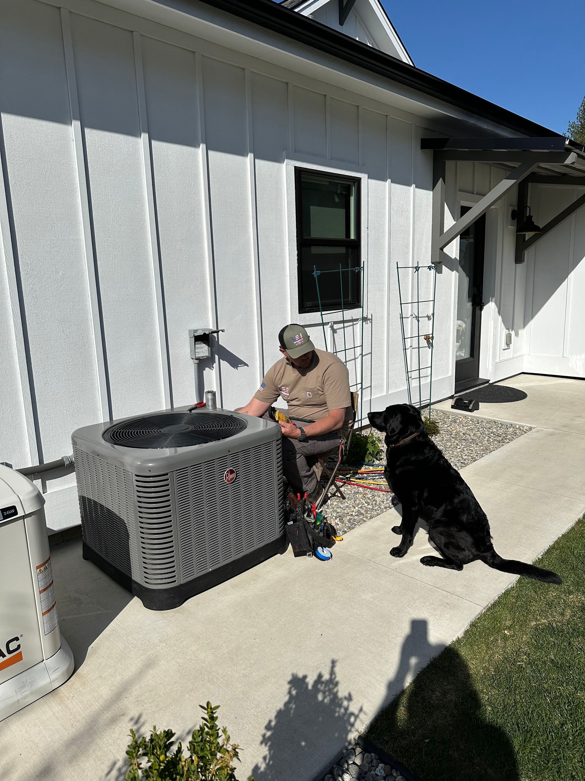 Man working on an AC unit outside, black dog sits nearby on a sunny day.