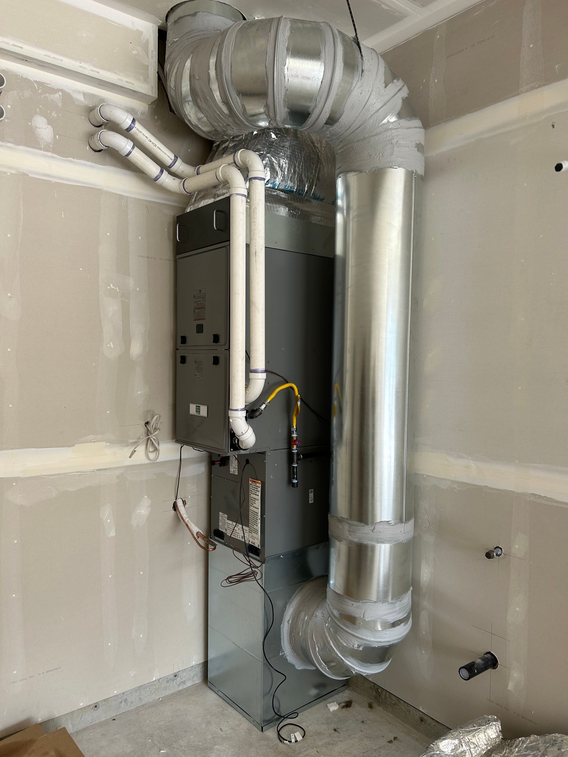 HVAC furnace system installed in a room with unfinished drywall. Silver ductwork and pipes are visible.