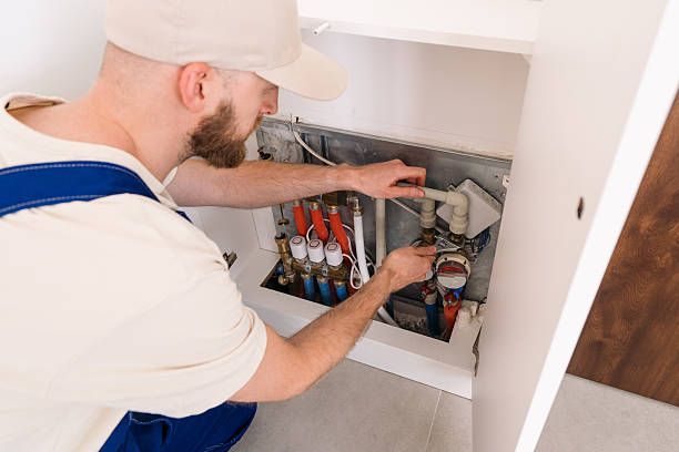 Plumber in a light beige uniform and cap works on pipes inside a white cabinet.